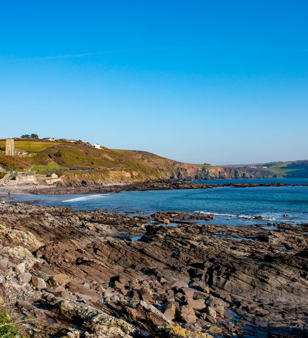 A rocky coastline with waves gently lapping at the shore, a small sandy beach, and a hill with a historic stone tower and scattered houses under a clear blue sky.