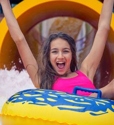 A smiling girl in a pink swimsuit rides a yellow inflatable tube down a water slide, raising her arms in excitement as water splashes around her.