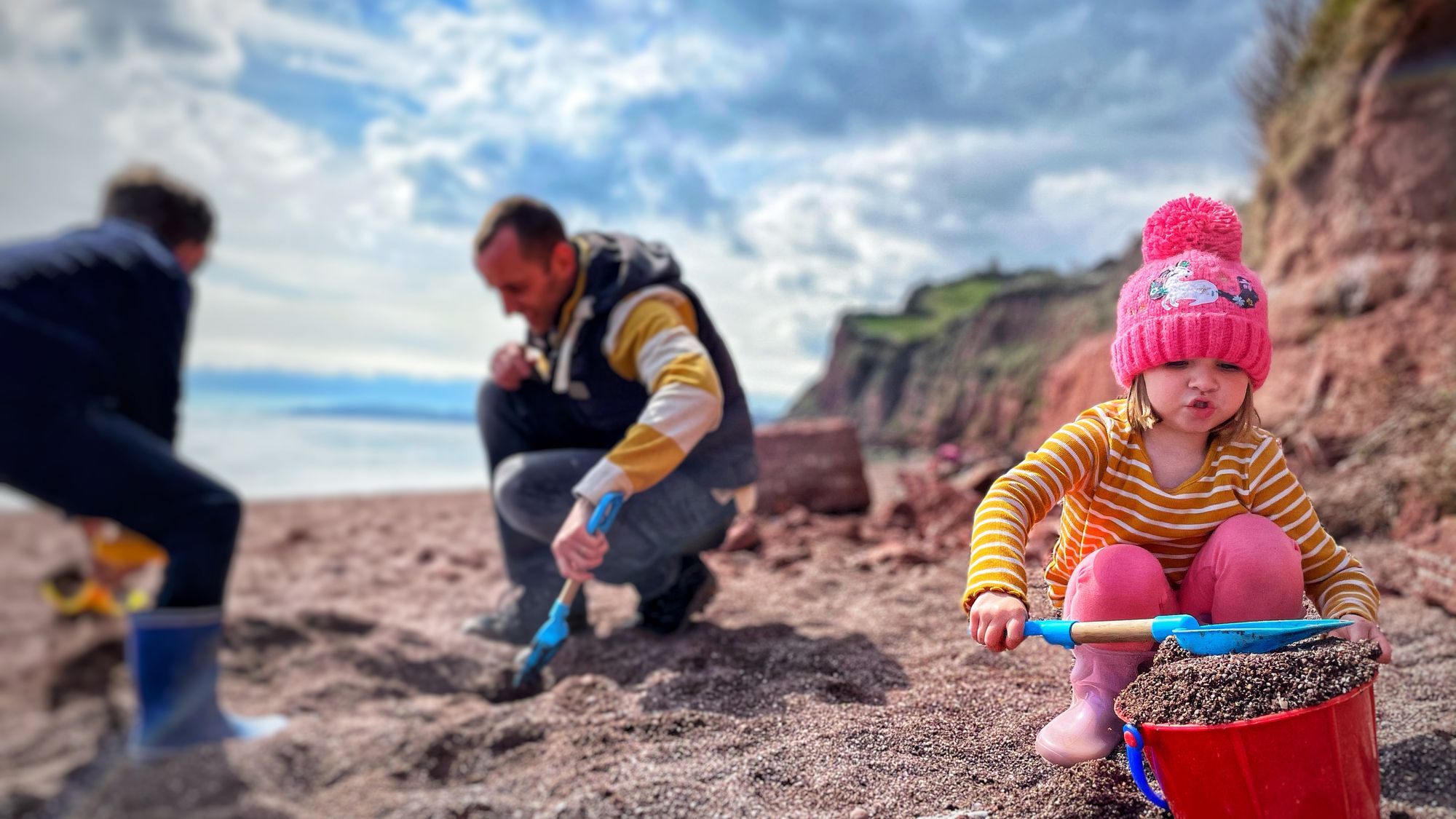A young child in a pink hat and striped shirt scoops sand into a red bucket on a beach, while two adults dig nearby. Cliffs and a cloudy sky are in the background.