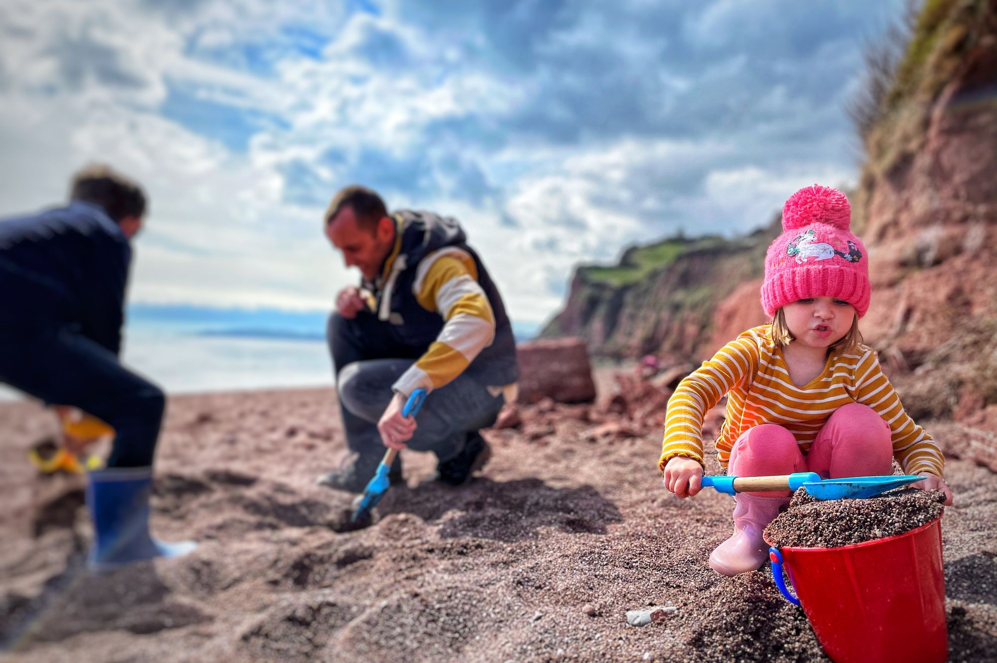 A young child in a pink hat and striped shirt scoops sand into a red bucket on a beach, while two adults dig nearby. Cliffs and a cloudy sky are in the background.