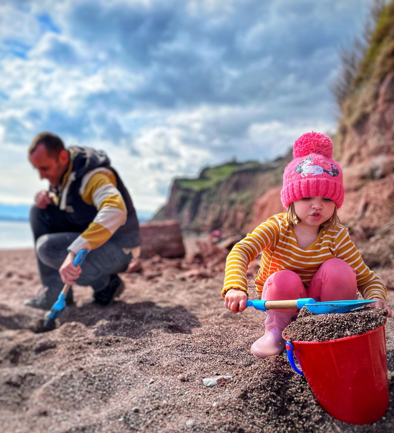 A young child in a pink hat and striped shirt scoops sand into a red bucket on a beach, while two adults dig nearby. Cliffs and a cloudy sky are in the background.