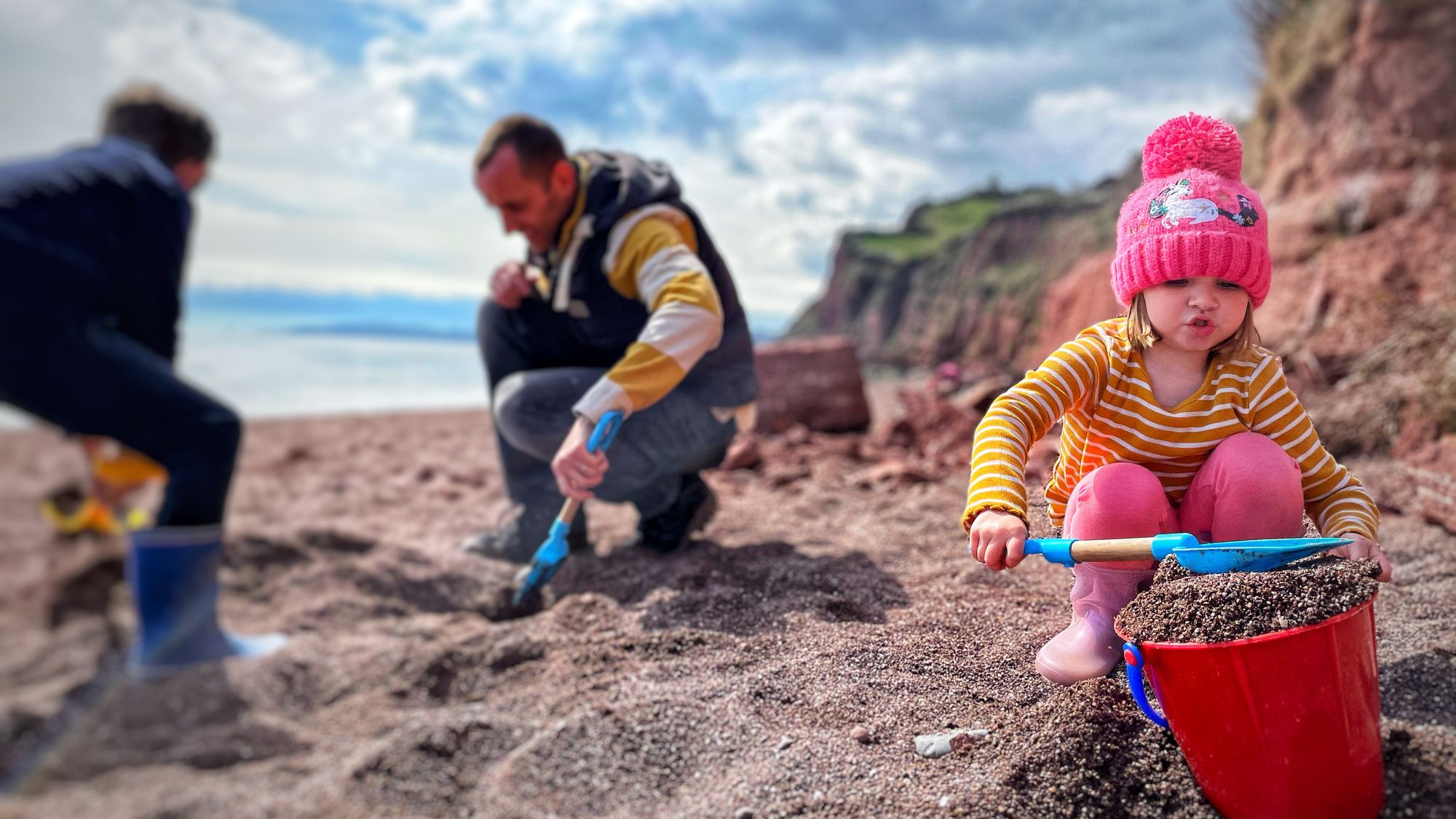 A young child in a pink hat and striped shirt scoops sand into a red bucket on a beach, while two adults dig nearby. Cliffs and a cloudy sky are in the background.