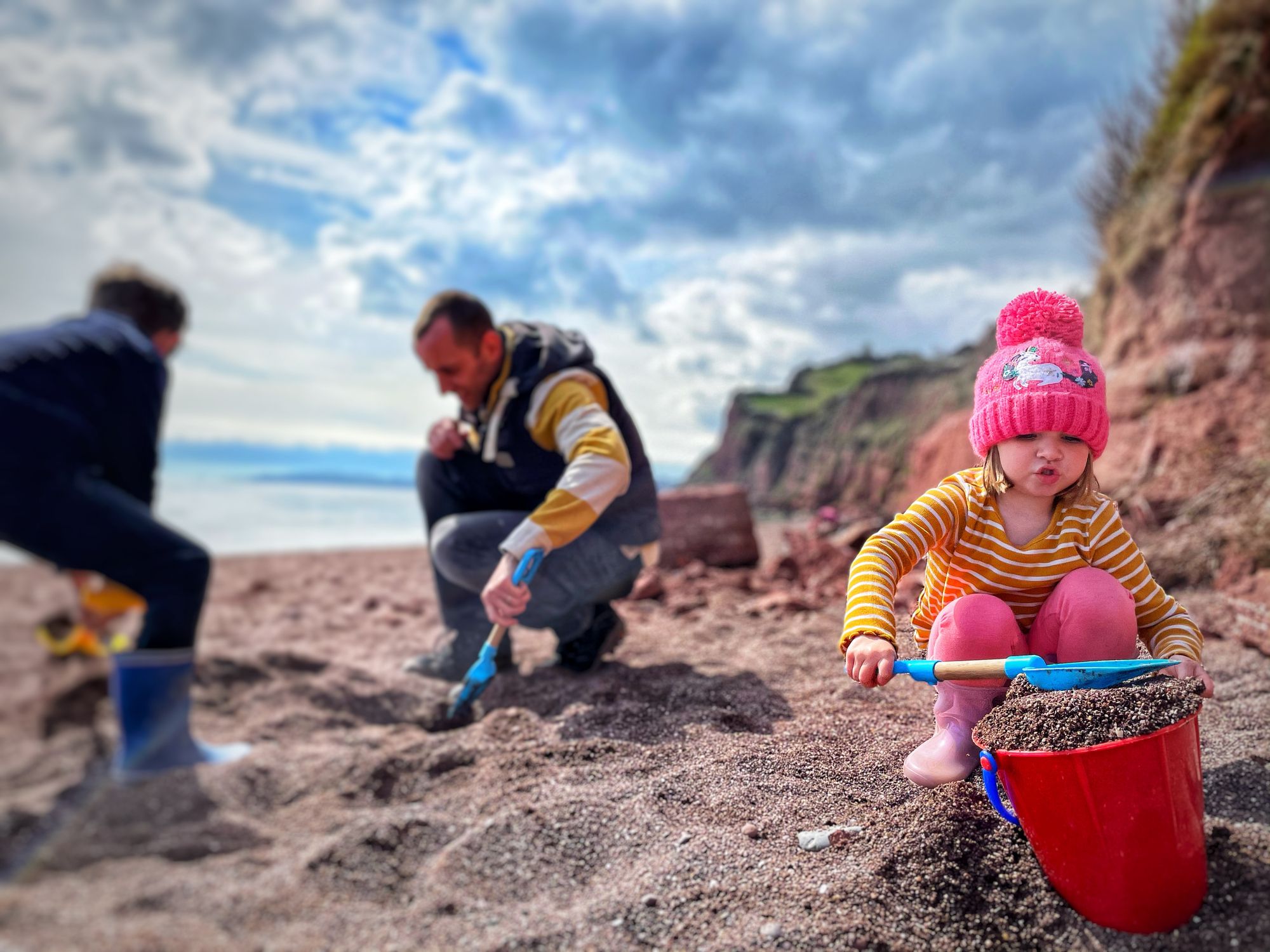 A young child in a pink hat and striped shirt scoops sand into a red bucket on a beach, while two adults dig nearby. Cliffs and a cloudy sky are in the background.