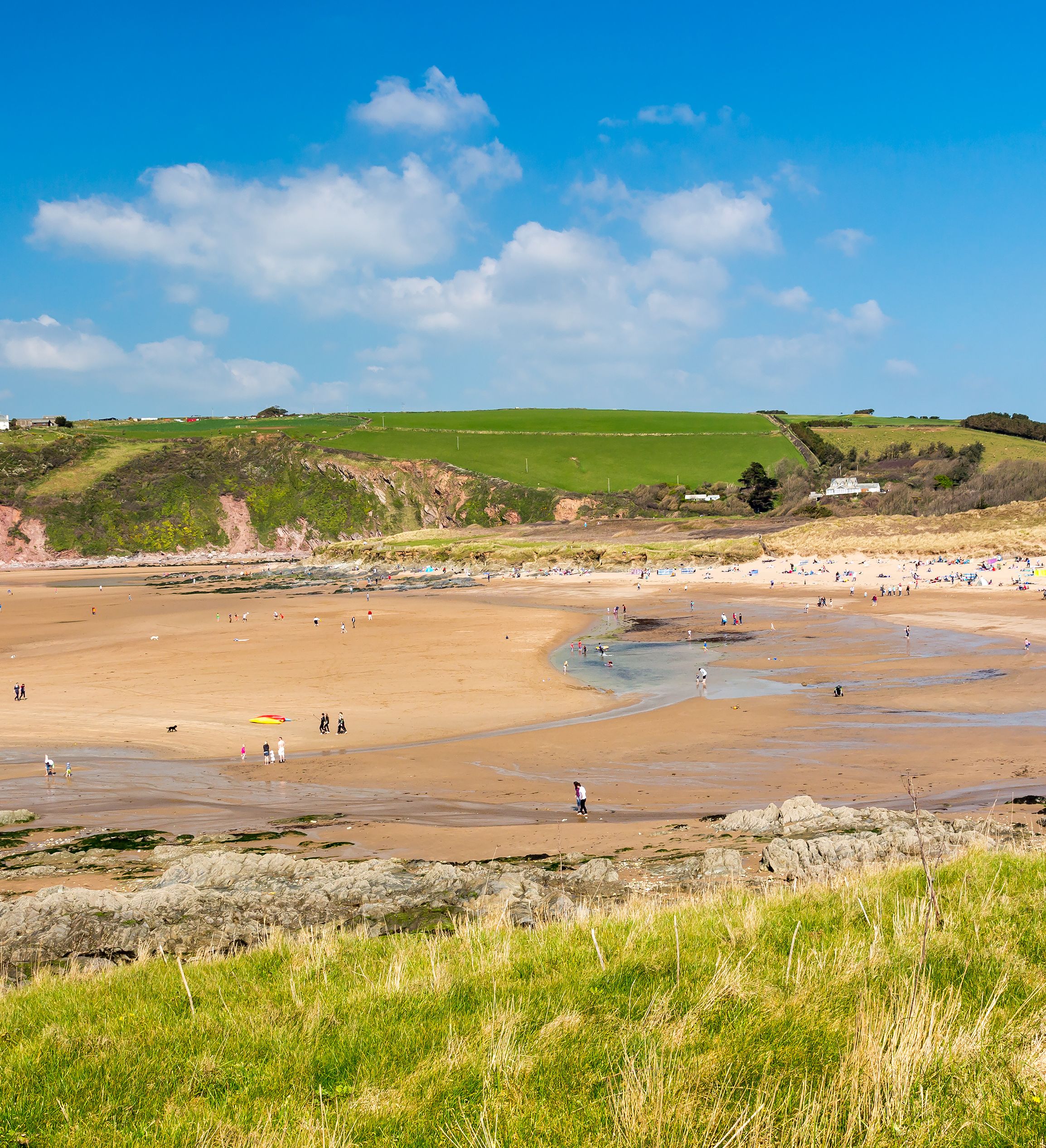 A wide sandy beach with people walking and relaxing stretches along a coastline backed by green rolling hills and cliffs under a bright blue sky with scattered clouds.