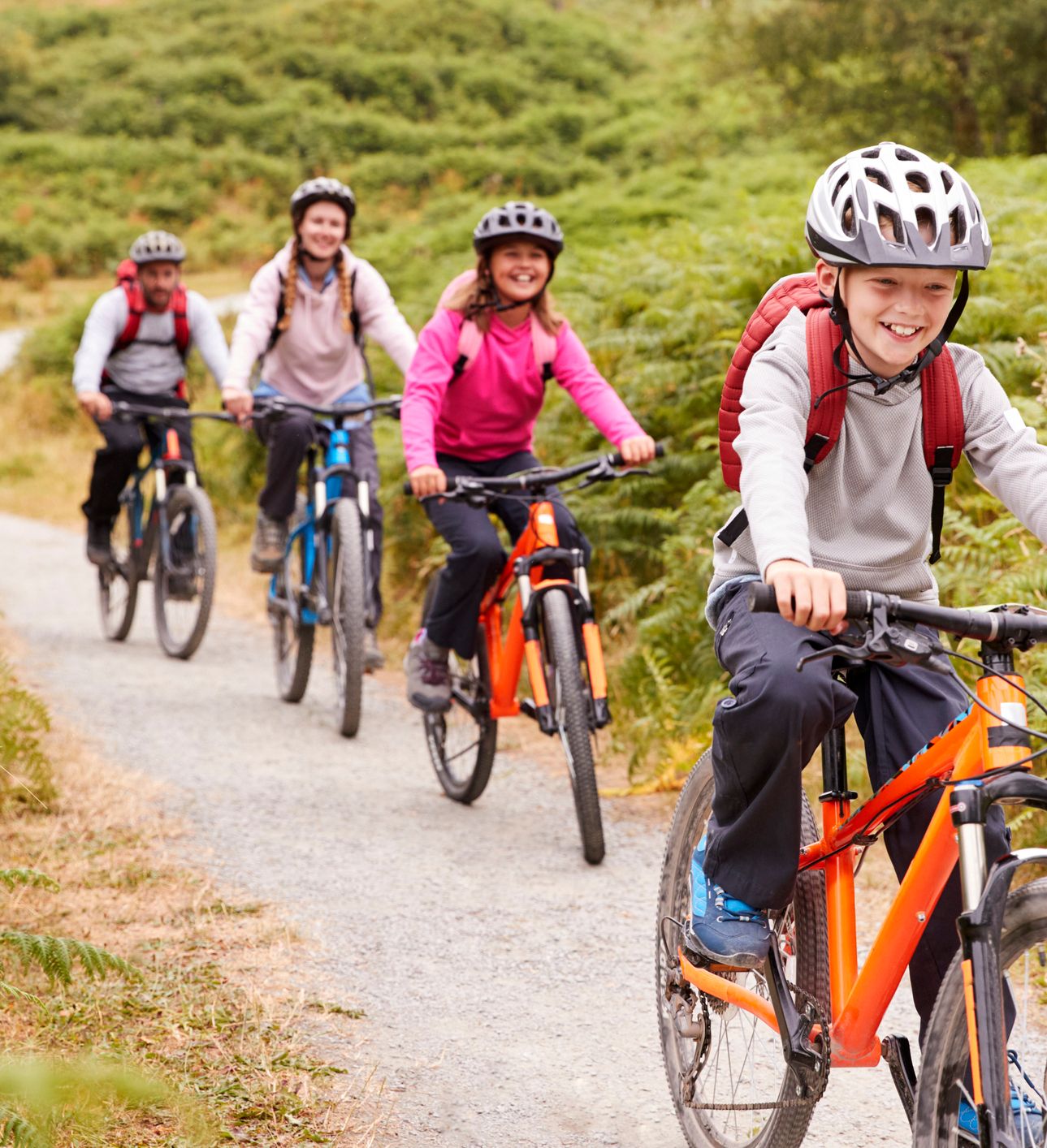 A group of four smiling cyclists, including children and adults, ride mountain bikes along a winding gravel trail surrounded by lush green ferns and rolling hills.