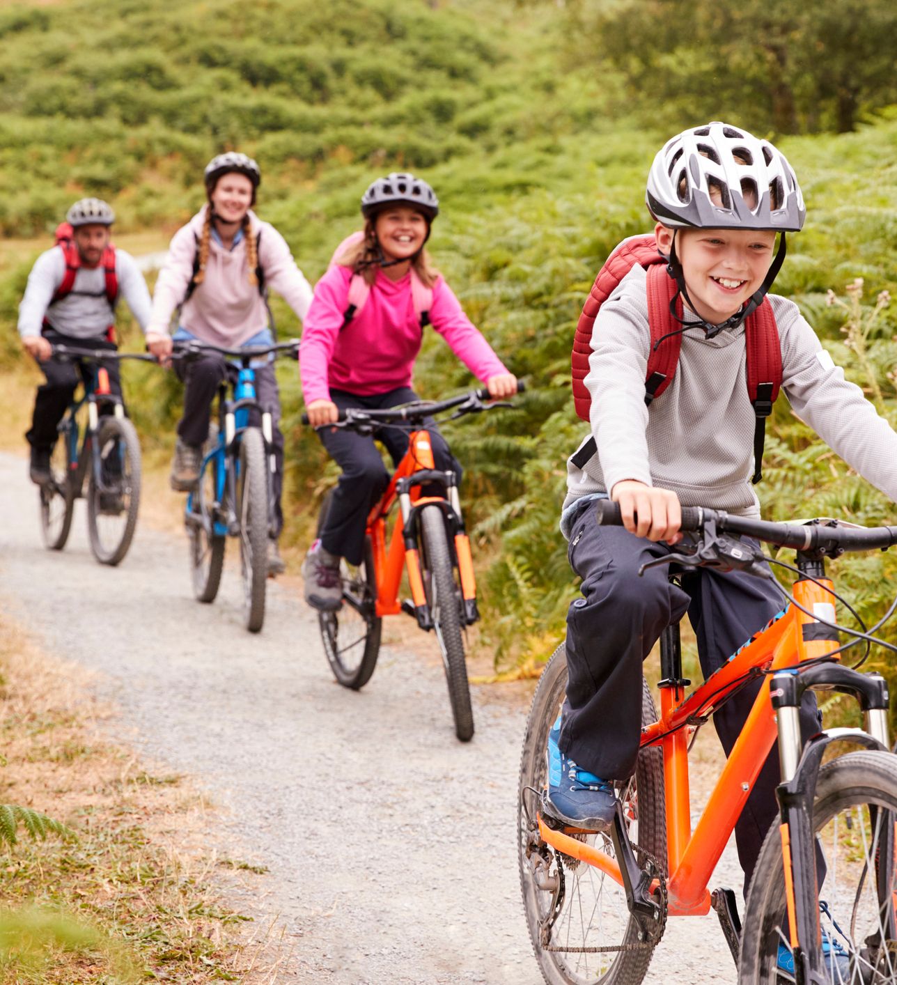 A group of four smiling cyclists, including children and adults, ride mountain bikes along a winding gravel trail surrounded by lush green ferns and rolling hills.