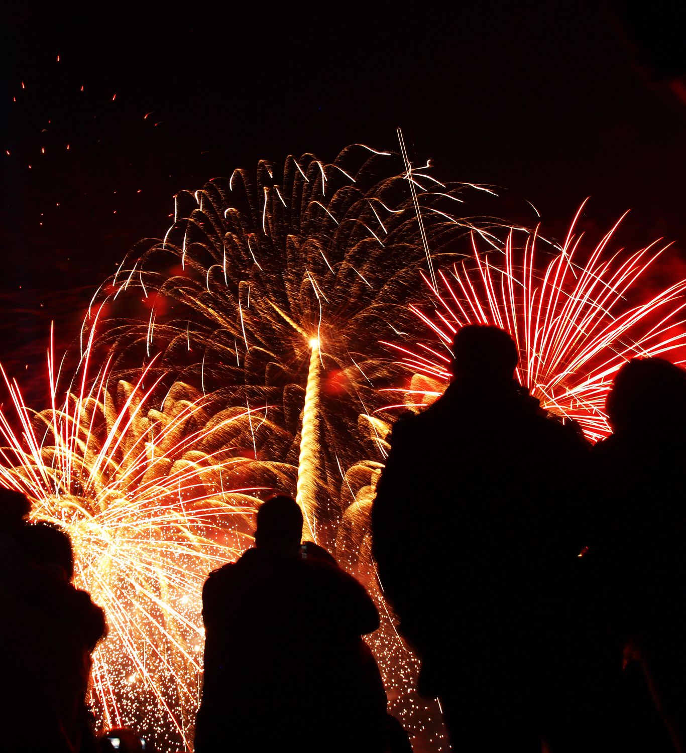 Silhouetted spectators watch a vibrant fireworks display light up the night sky with bursts of red, gold, and orange, creating a dazzling celebration scene.