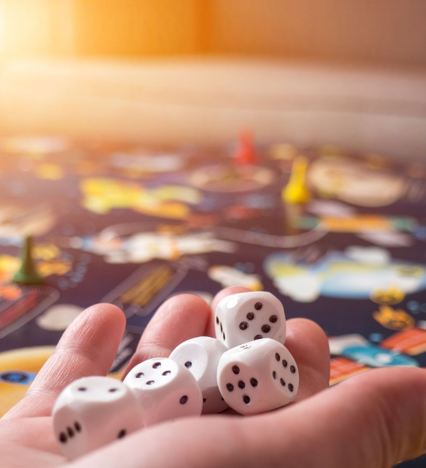 A hand holds white dice over a colorful space-themed board game. Game pieces are scattered across the board, with warm sunlight casting a glow over the scene.