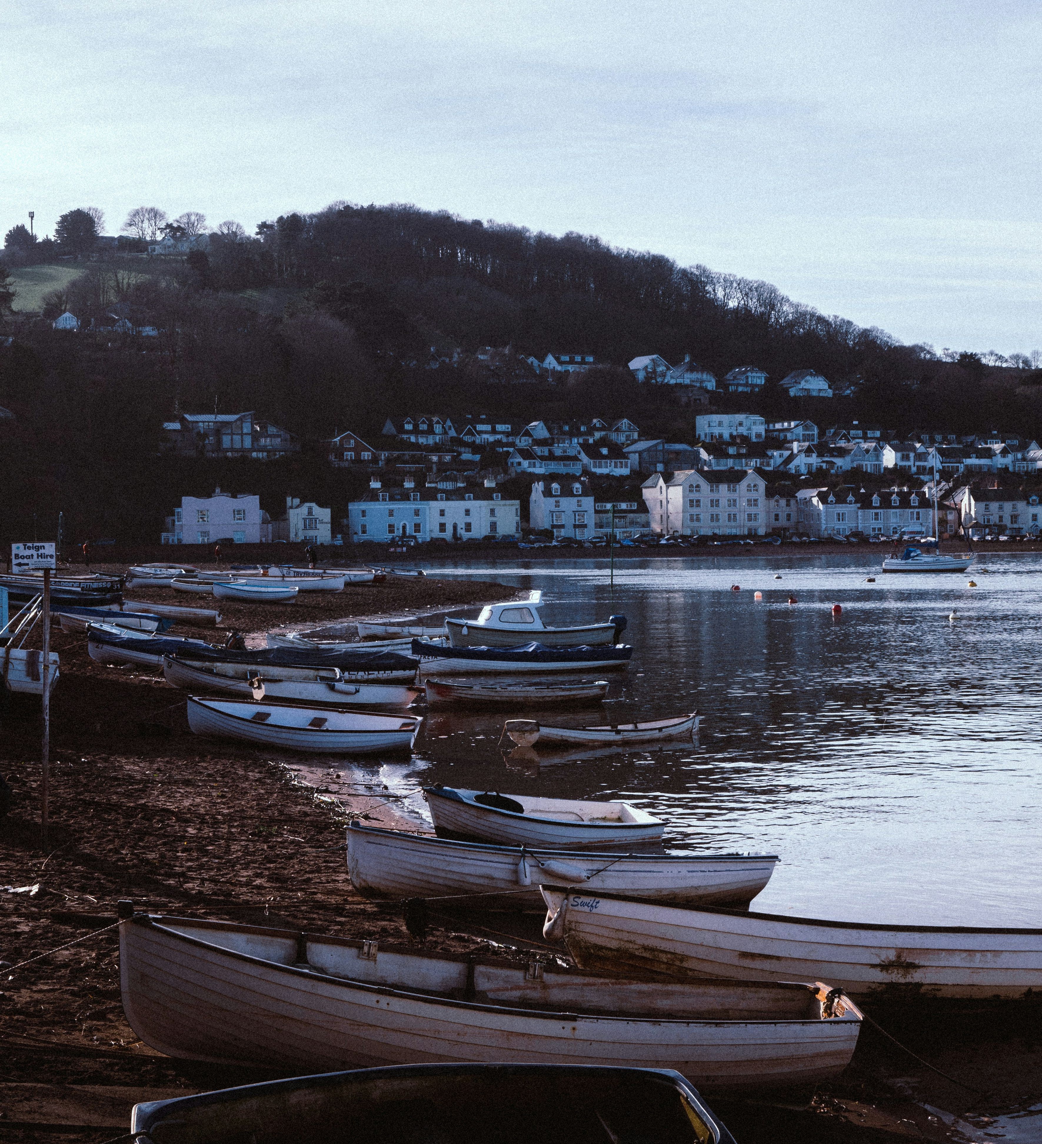 Small boats on the shore by a calm harbor with a hillside village in the background