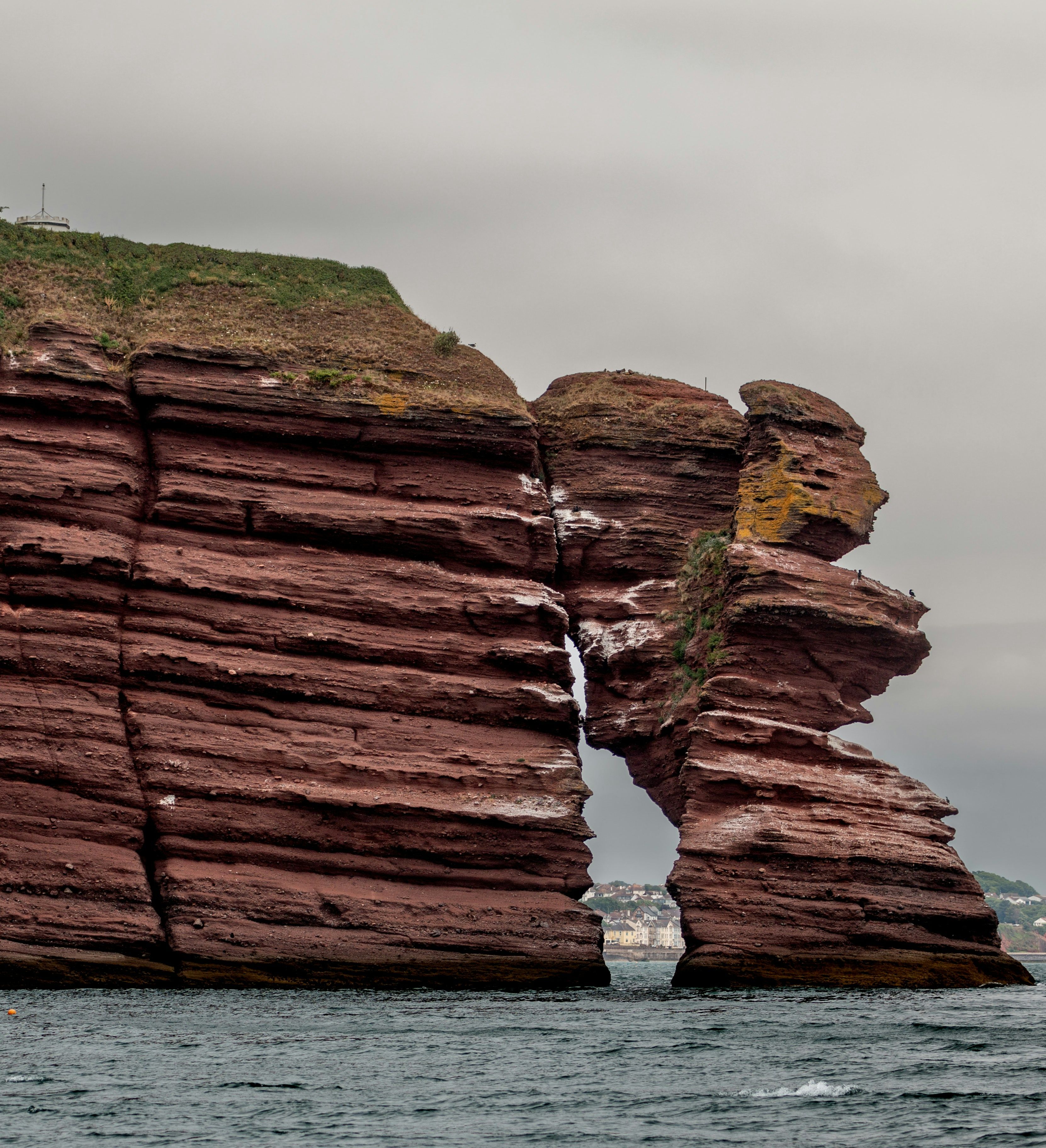 Eroded red sandstone cliffs with an arch formation on the coast, viewed from the water under a cloudy sky.