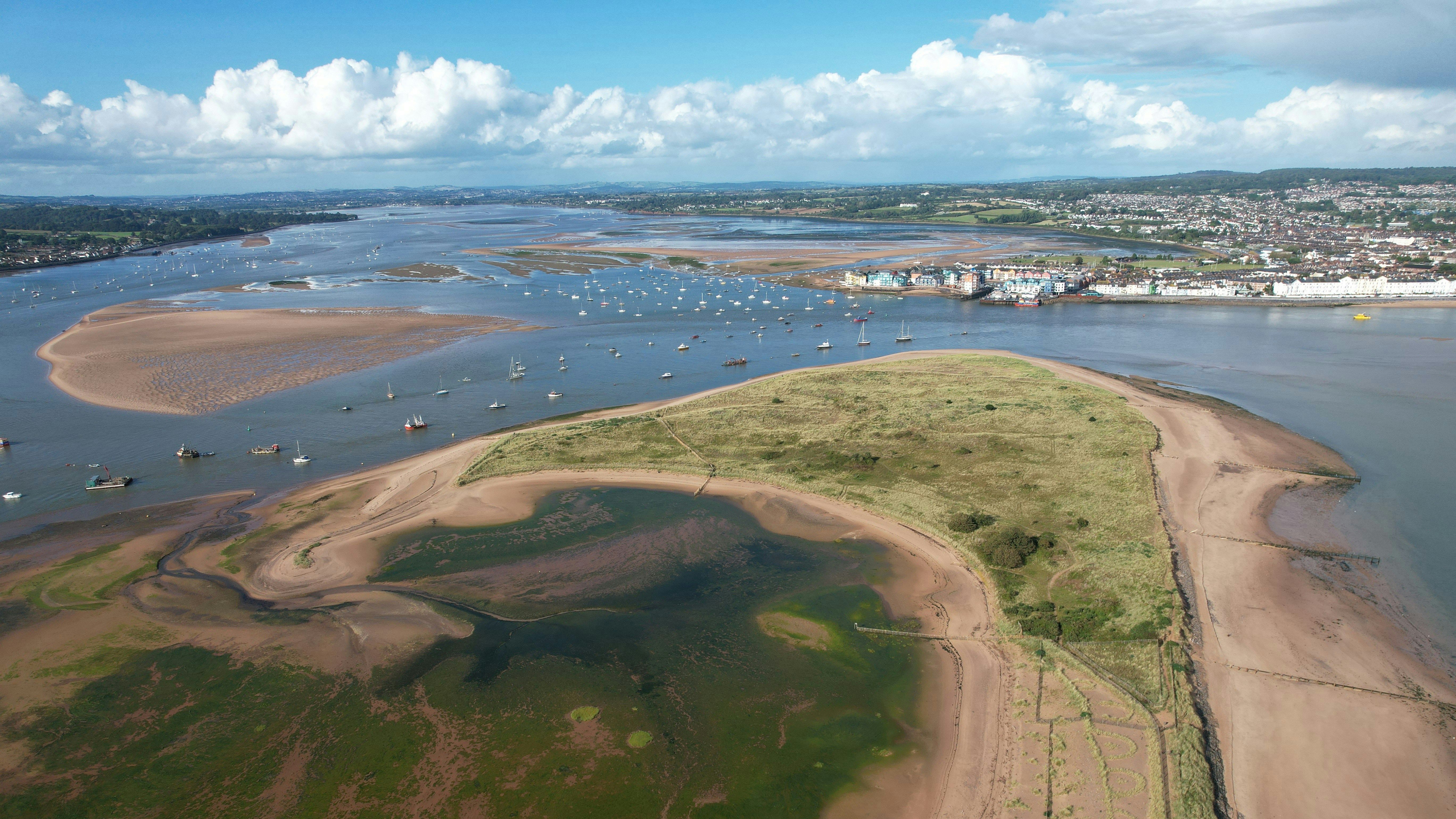 Aerial view of a coastal sandbank with grassy patches, boats anchored in the water, and a town in the distance under a cloudy blue sky.
