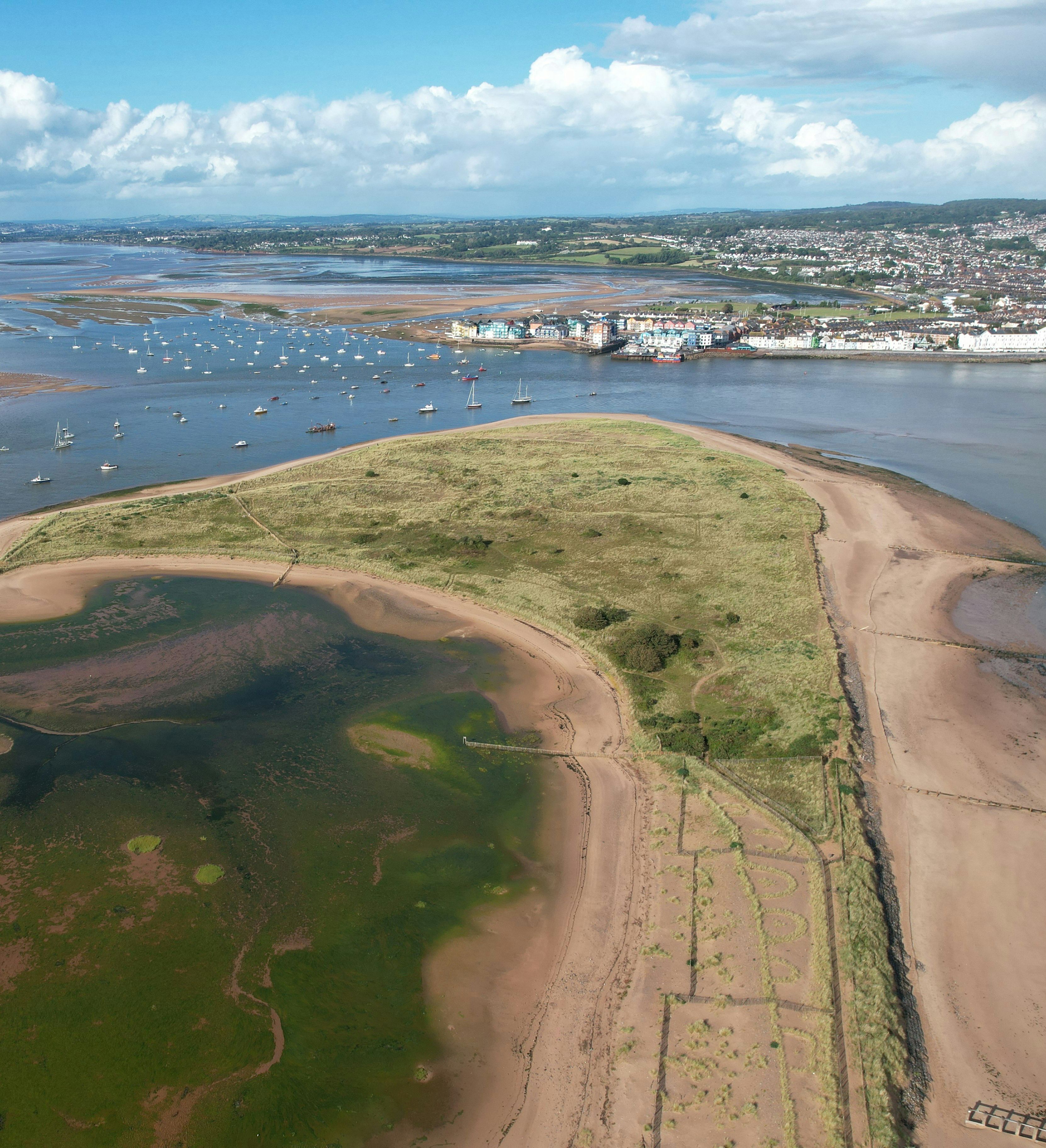 Aerial view of a coastal sandbank with grassy patches, boats anchored in the water, and a town in the distance under a cloudy blue sky.