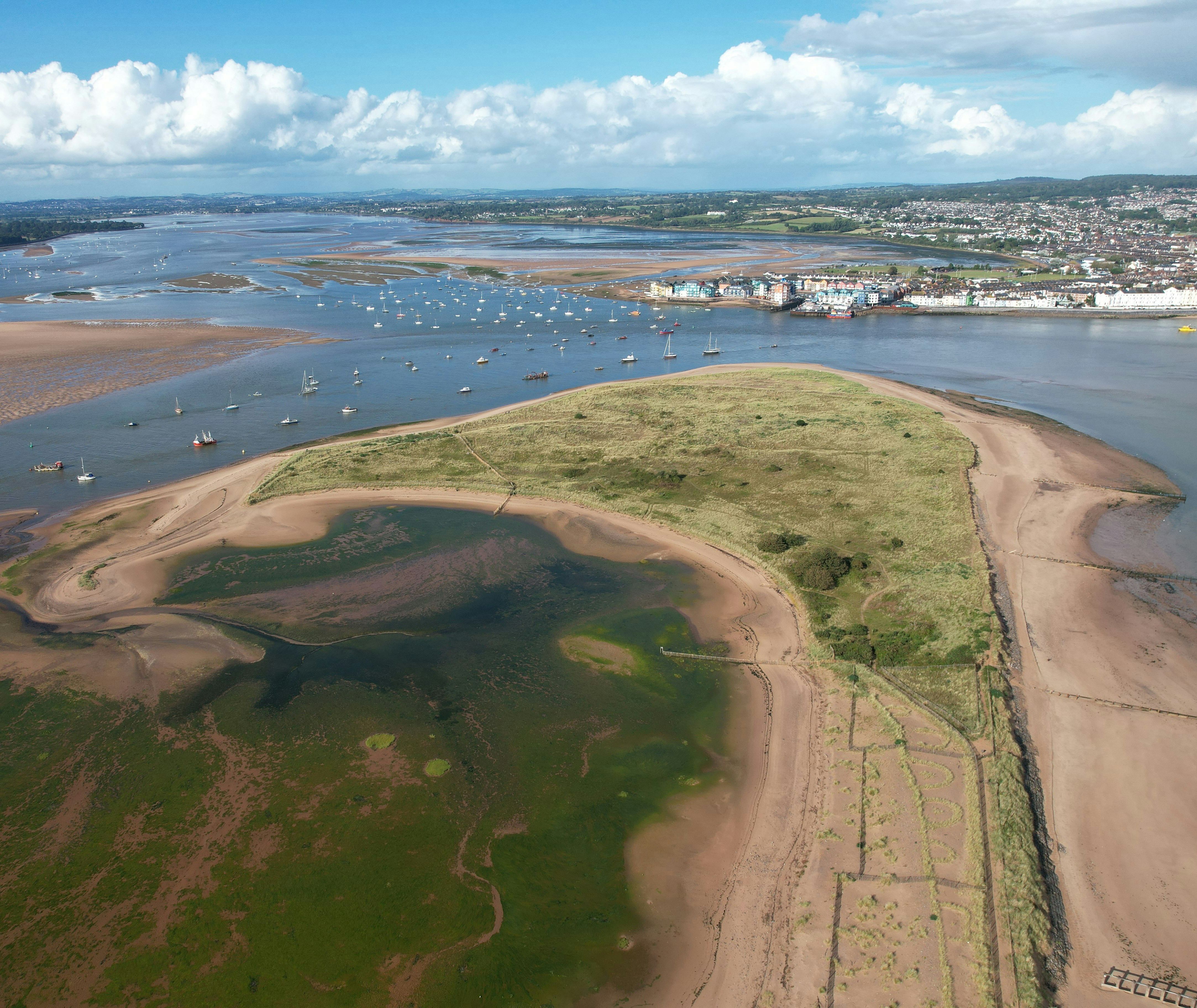 Aerial view of a coastal sandbank with grassy patches, boats anchored in the water, and a town in the distance under a cloudy blue sky.