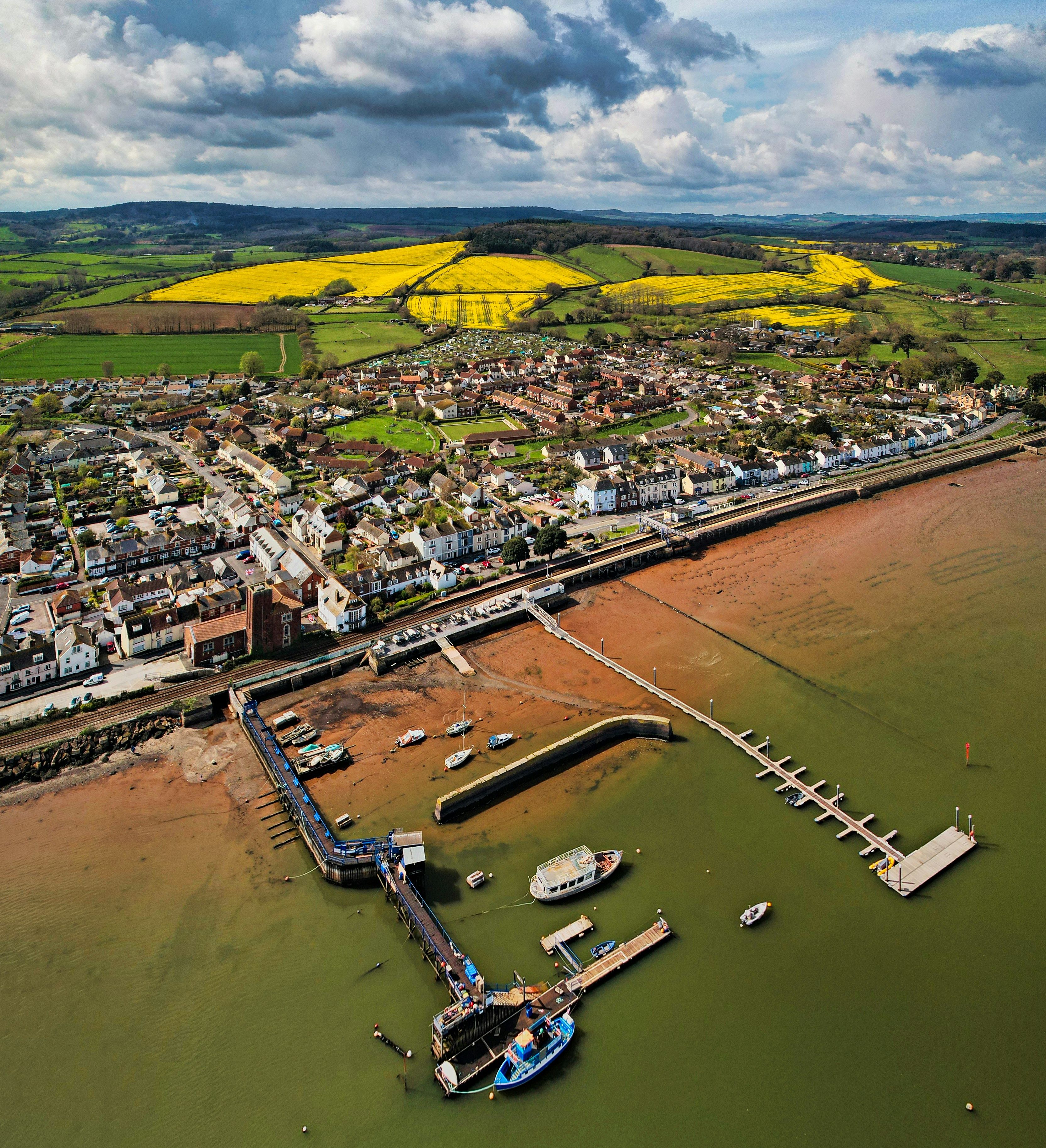 Aerial view of a coastal town with a marina, boats and colorful fields in the background.