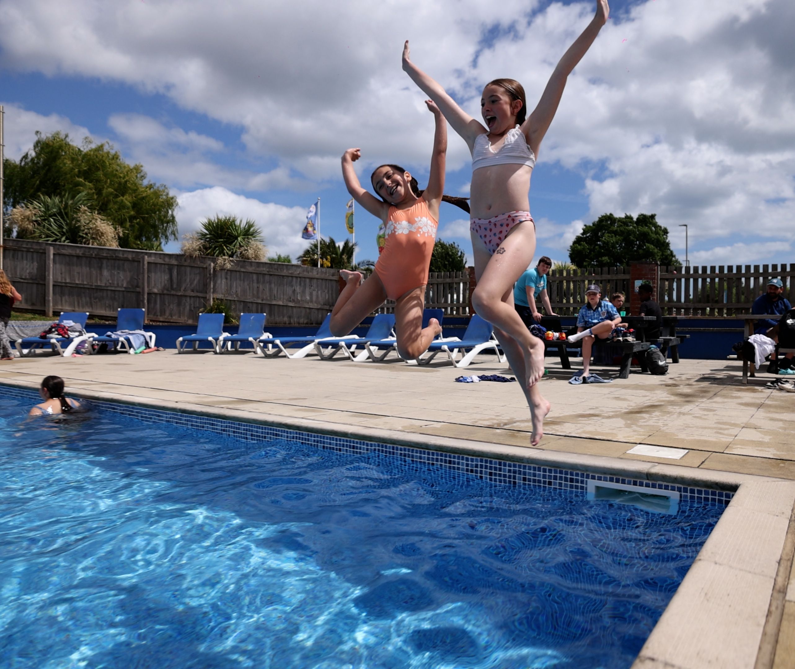 Two children in swimsuits joyfully jump into an outdoor swimming pool on a sunny day while others relax nearby.