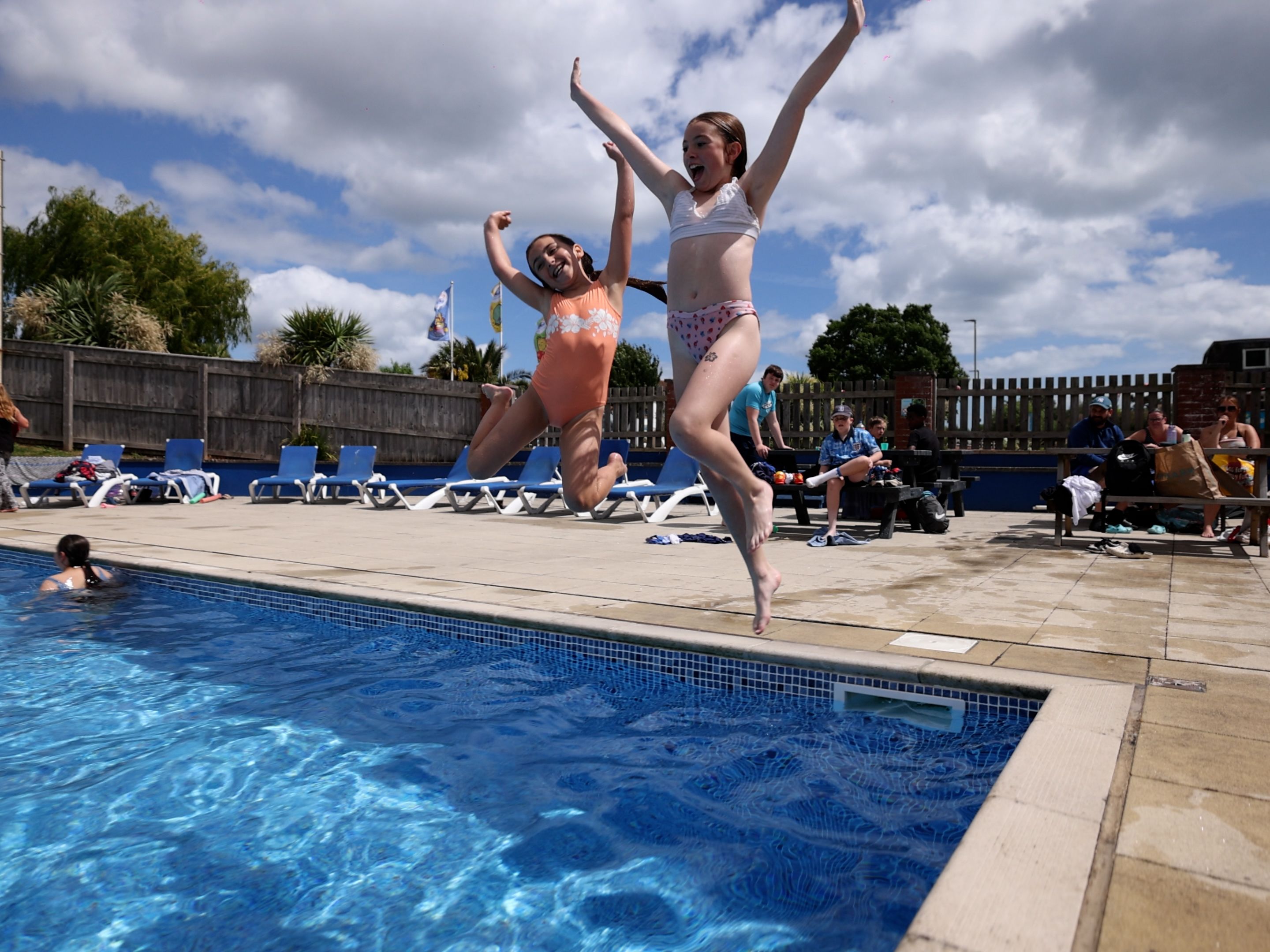Two children in swimsuits joyfully jump into an outdoor swimming pool on a sunny day while others relax nearby.