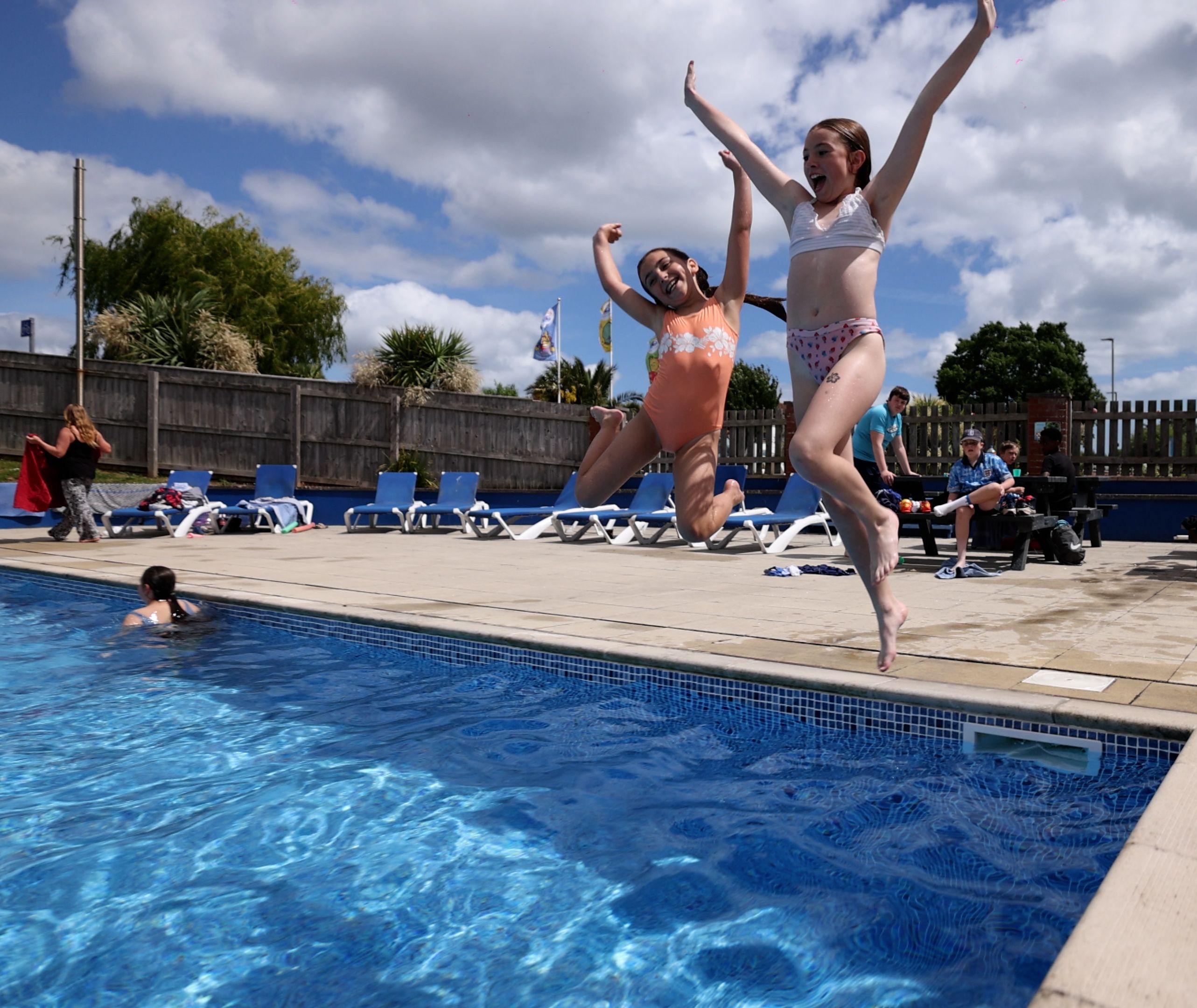 Two children in swimsuits joyfully jump into an outdoor swimming pool on a sunny day while others relax nearby.