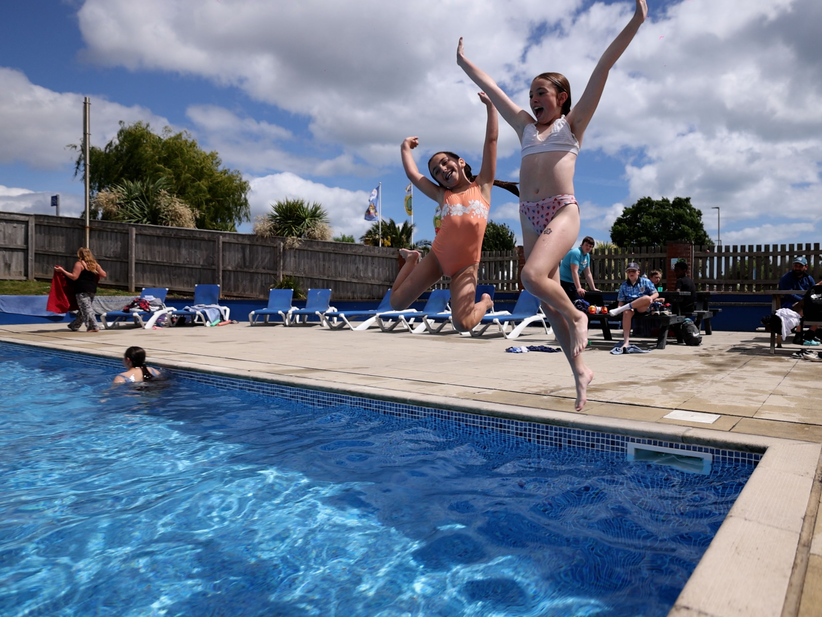Two children in swimsuits joyfully jump into an outdoor swimming pool on a sunny day while others relax nearby.