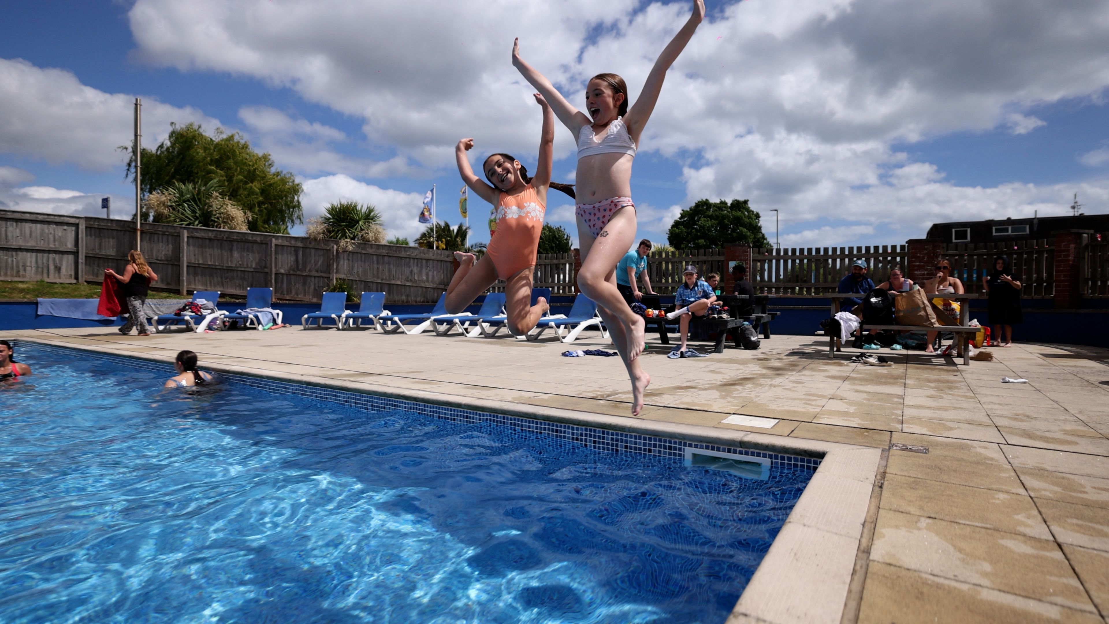 Two children in swimsuits joyfully jump into an outdoor swimming pool on a sunny day while others relax nearby.