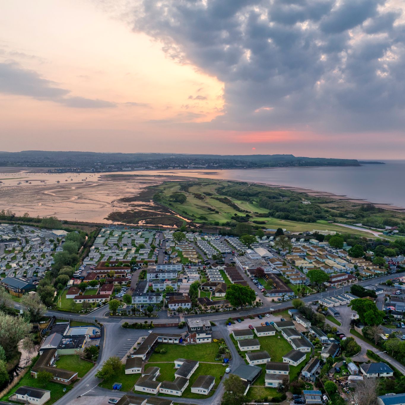 Aerial view of a coastal town with residential areas, green fields, and the sea at sunset under a cloudy sky.