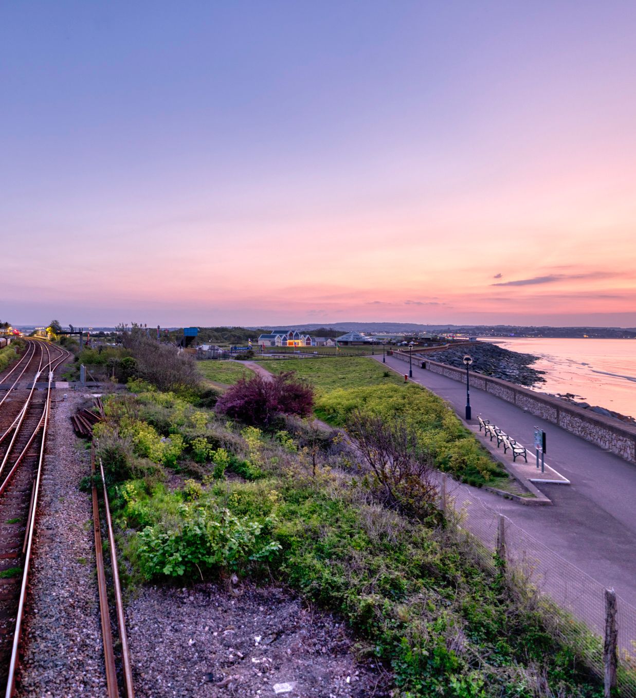 Railway tracks by the coast at sunset with a walking path and benches along the shoreline.