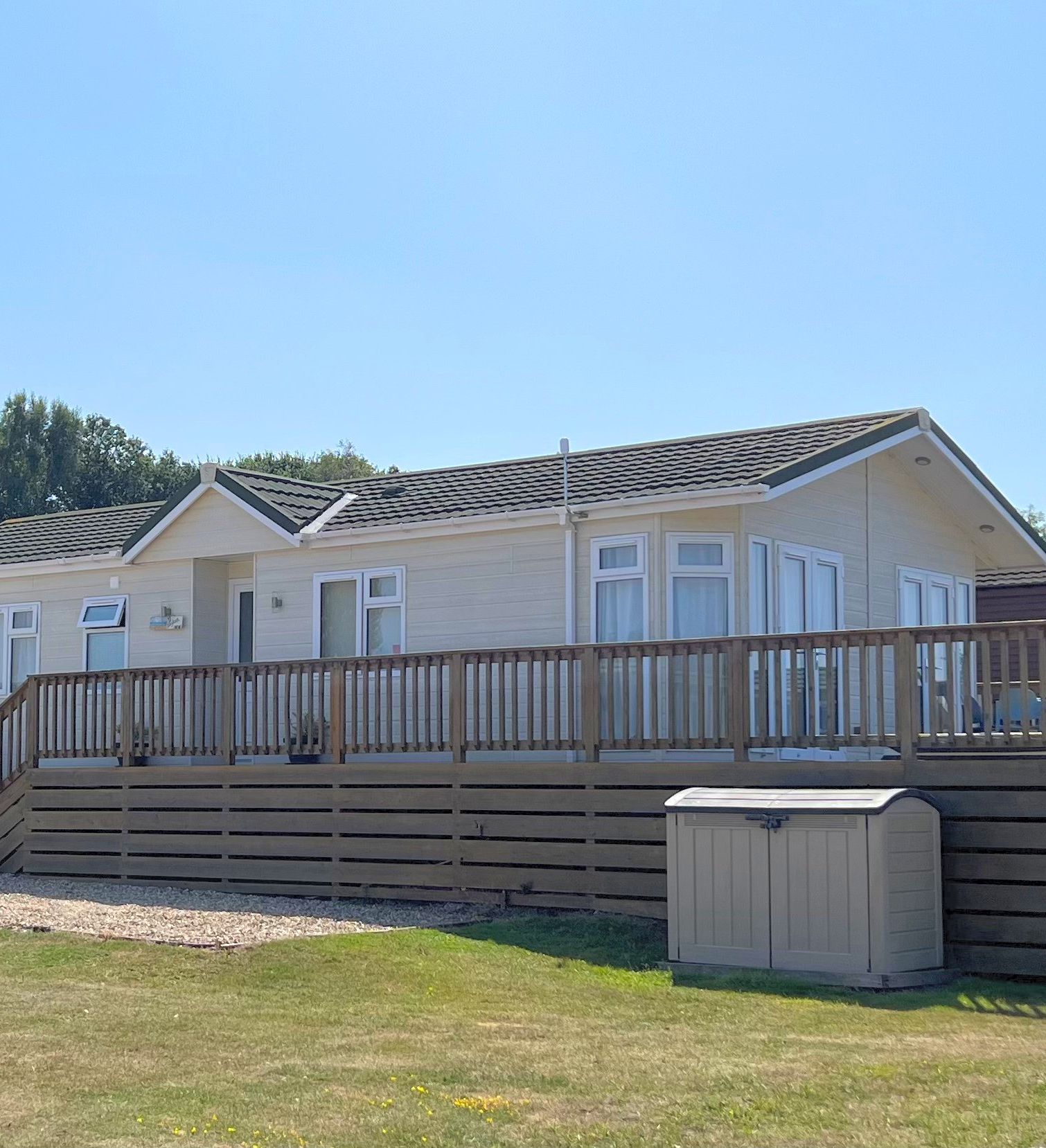 Exterior view of a single-story light-colored mobile home with a wooden deck and railing.
