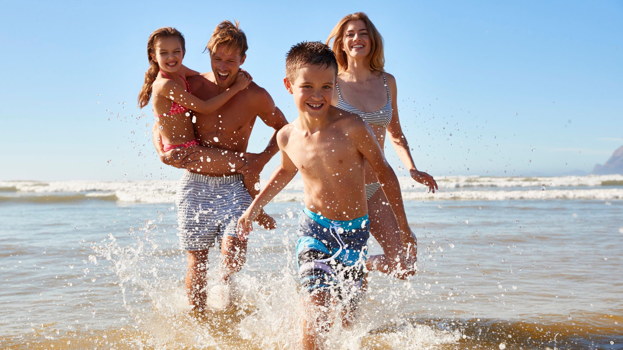 A happy family of four splashing and playing in shallow water at the beach on a sunny day.