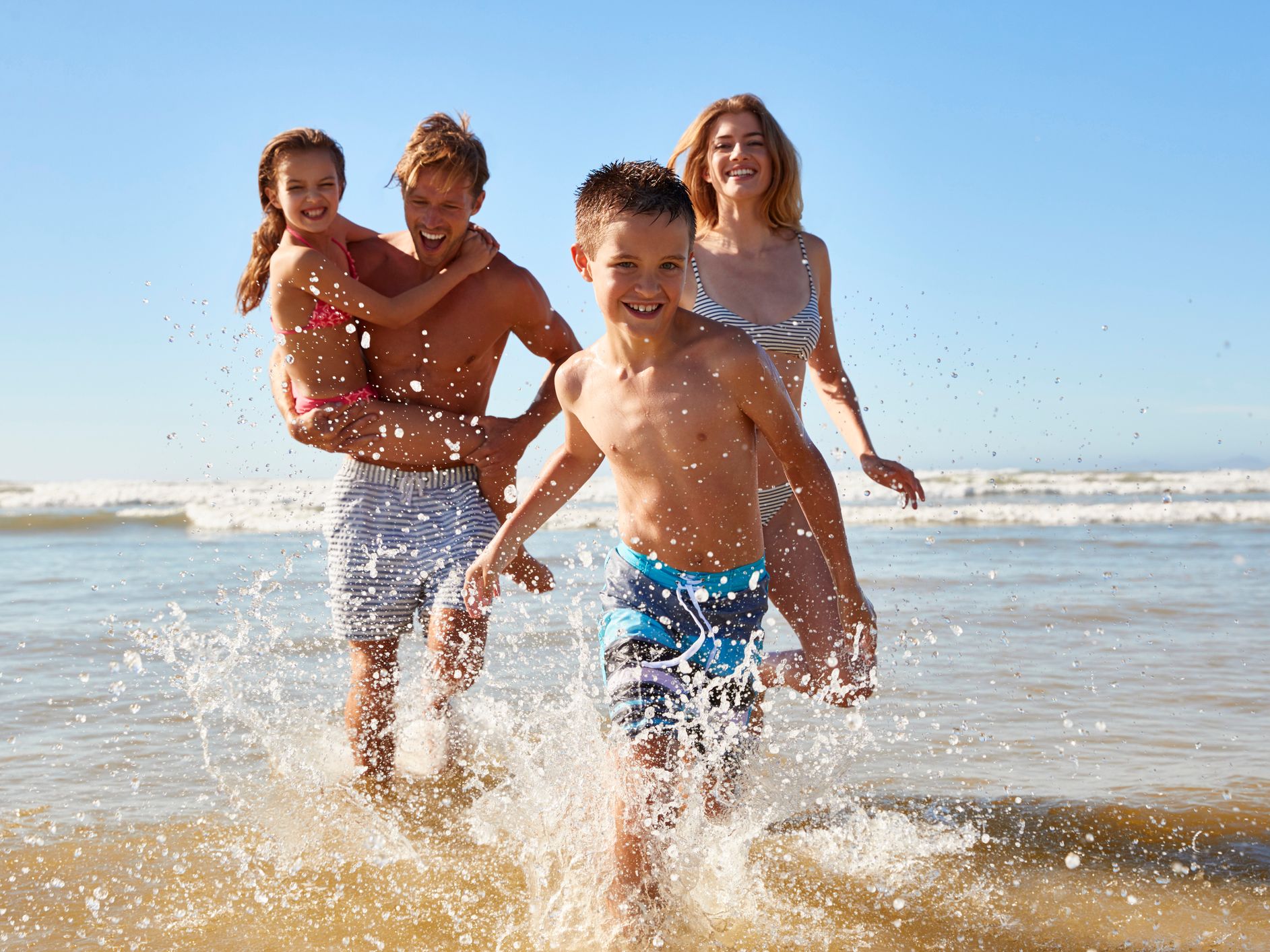 A happy family of four splashing and playing in shallow water at the beach on a sunny day.