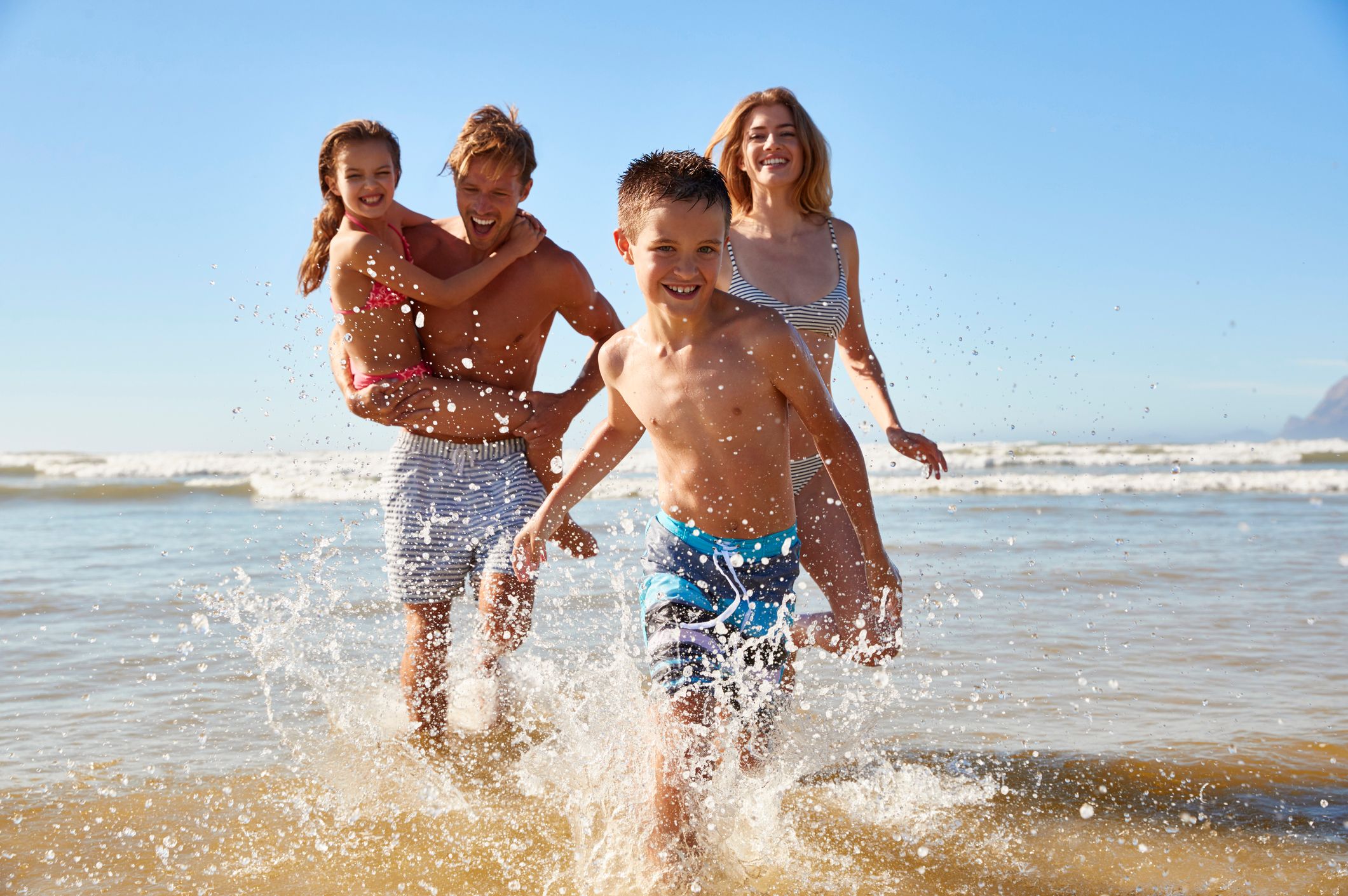 A happy family of four splashing and playing in shallow water at the beach on a sunny day.