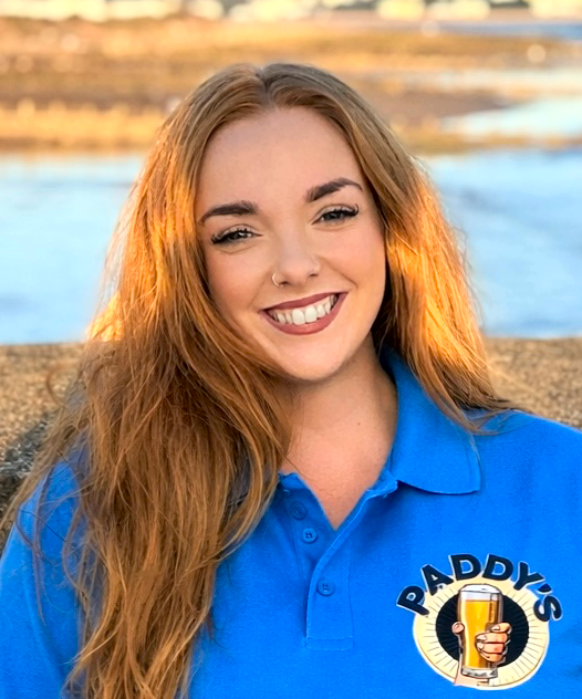 Smiling woman with long hair wearing a blue Paddy's shirt standing outdoors by the water