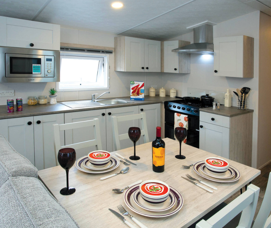 Modern kitchen-diner with white cabinets, built-in microwave, sink and cooker, and a table set with bowls, cutlery, wine glasses and a bottle of red wine.