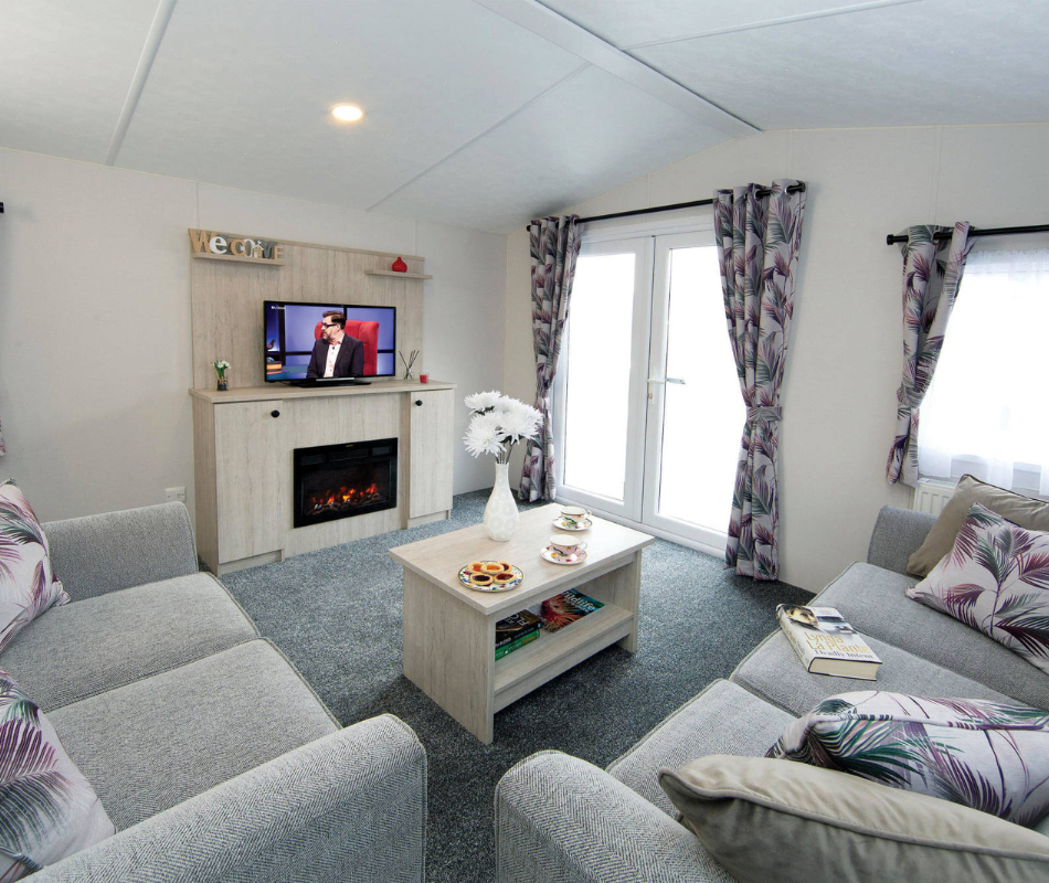 Bright lounge with grey sofas, coffee table, TV and electric fireplace, and patterned curtains by French doors