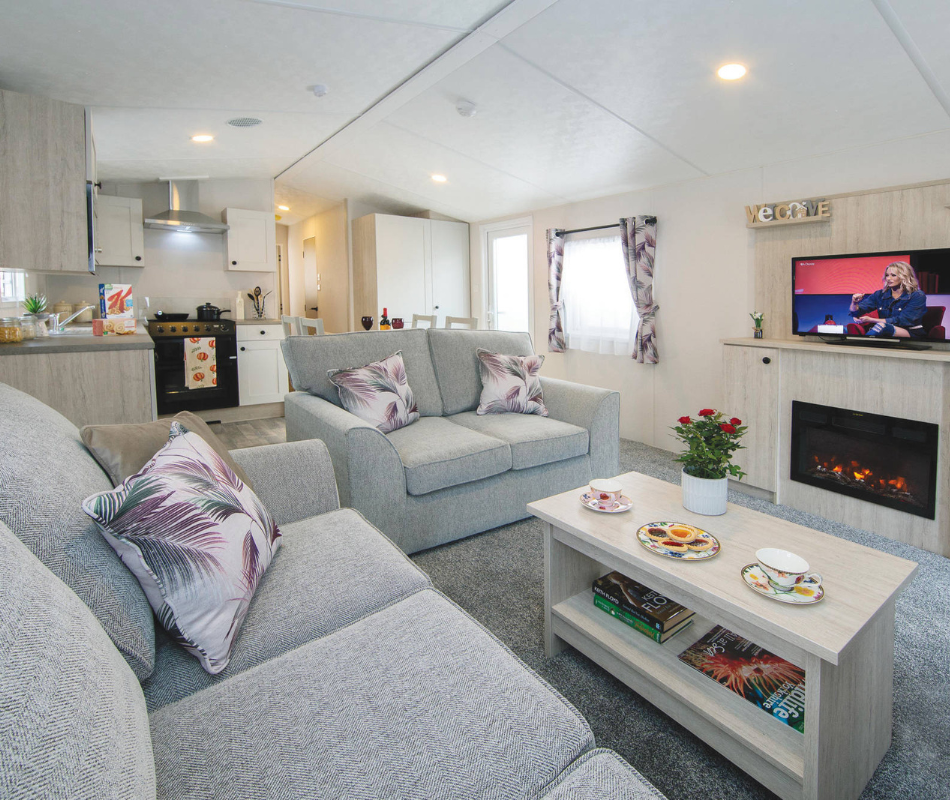 Bright open-plan living room with grey sofas, coffee table and kitchenette, with TV and electric fireplace