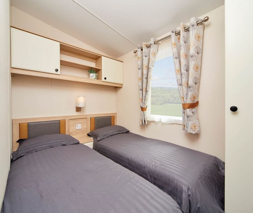 Compact twin bedroom with two single beds, overhead cupboards and window with patterned curtains