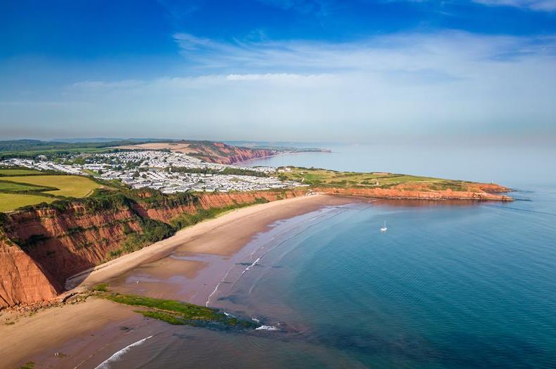 Aerial view of a sandy bay with red cliffs, calm sea and a coastal caravan park under a blue sky