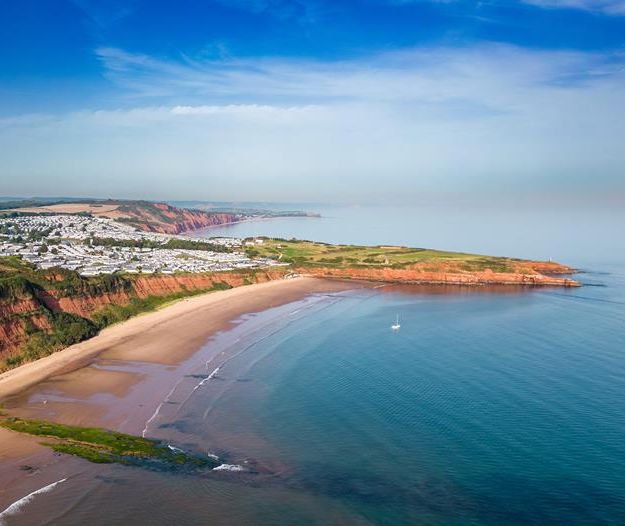 Aerial view of a sandy bay with red cliffs, calm sea and a coastal caravan park under a blue sky