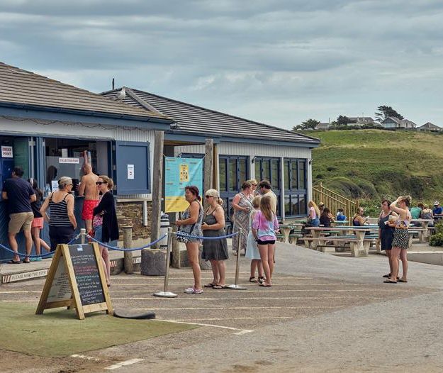 People queuing outside a seaside café by the beach with picnic tables and grassy cliffs in the background