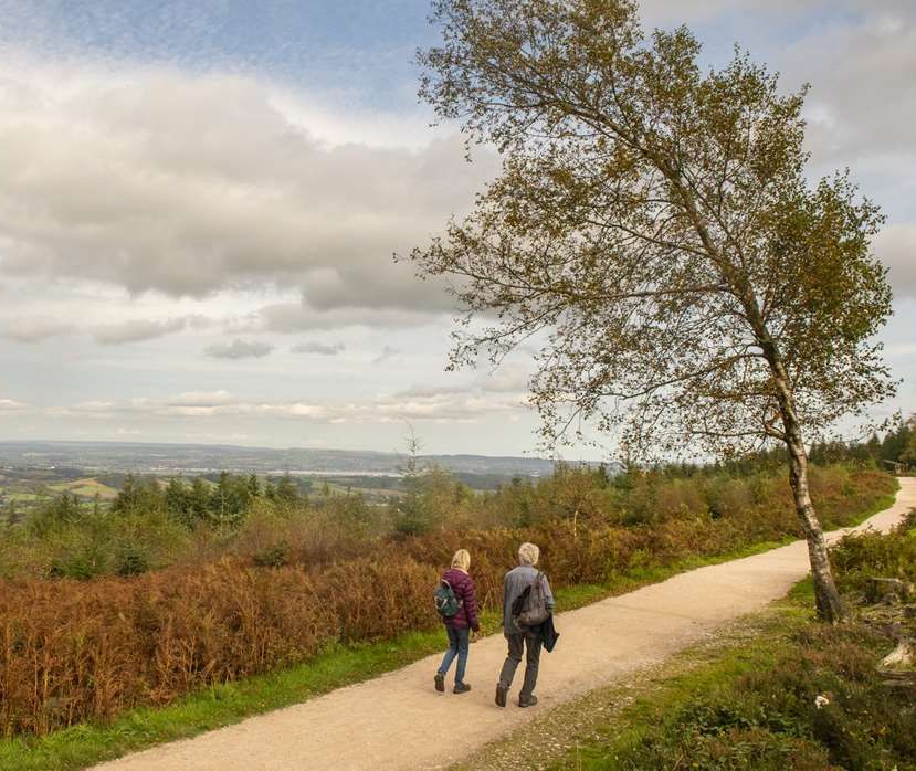 Two walkers on a woodland path overlooking countryside under a cloudy sky