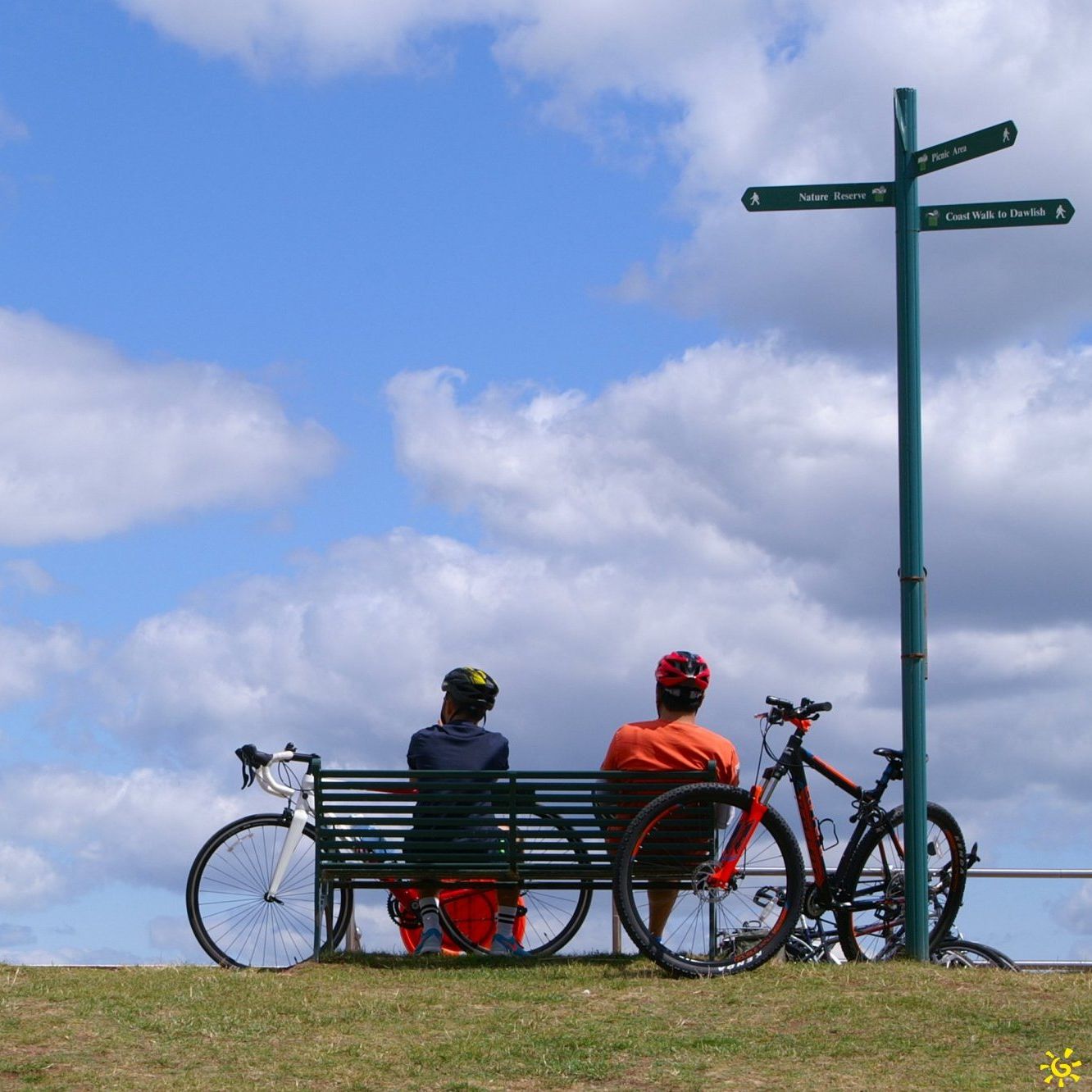 Two cyclists resting on a bench with their bicycles beside a signpost on the coast walk to Dawlish