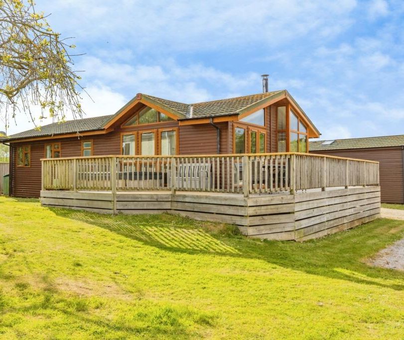 Modern wooden lodge with large windows and wraparound deck on a grassy lawn under a blue sky