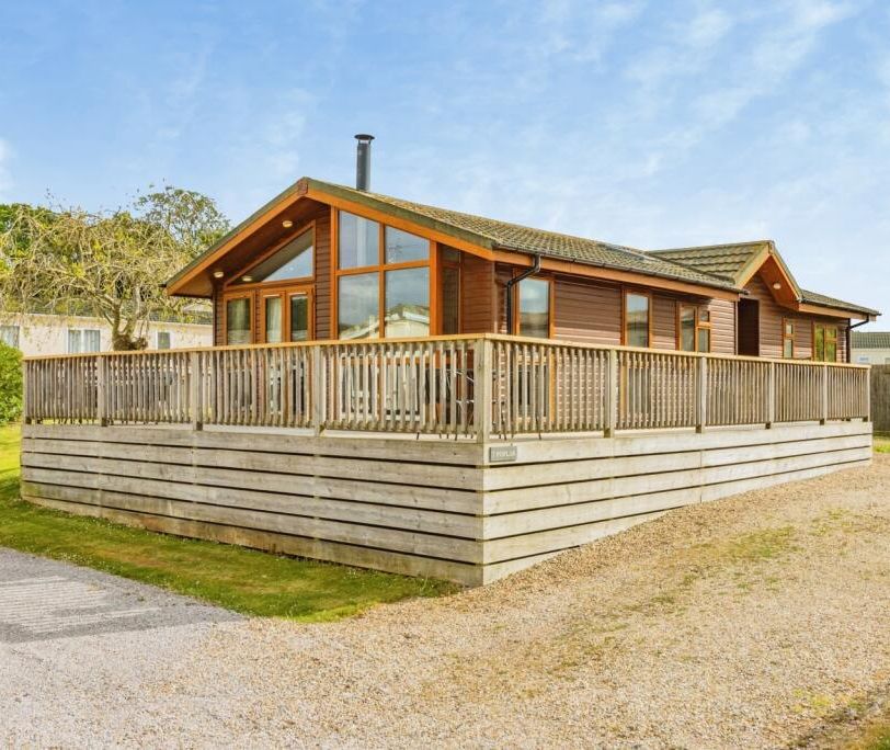Modern timber lodge with large windows and wraparound deck on a gravel plot under a blue sky