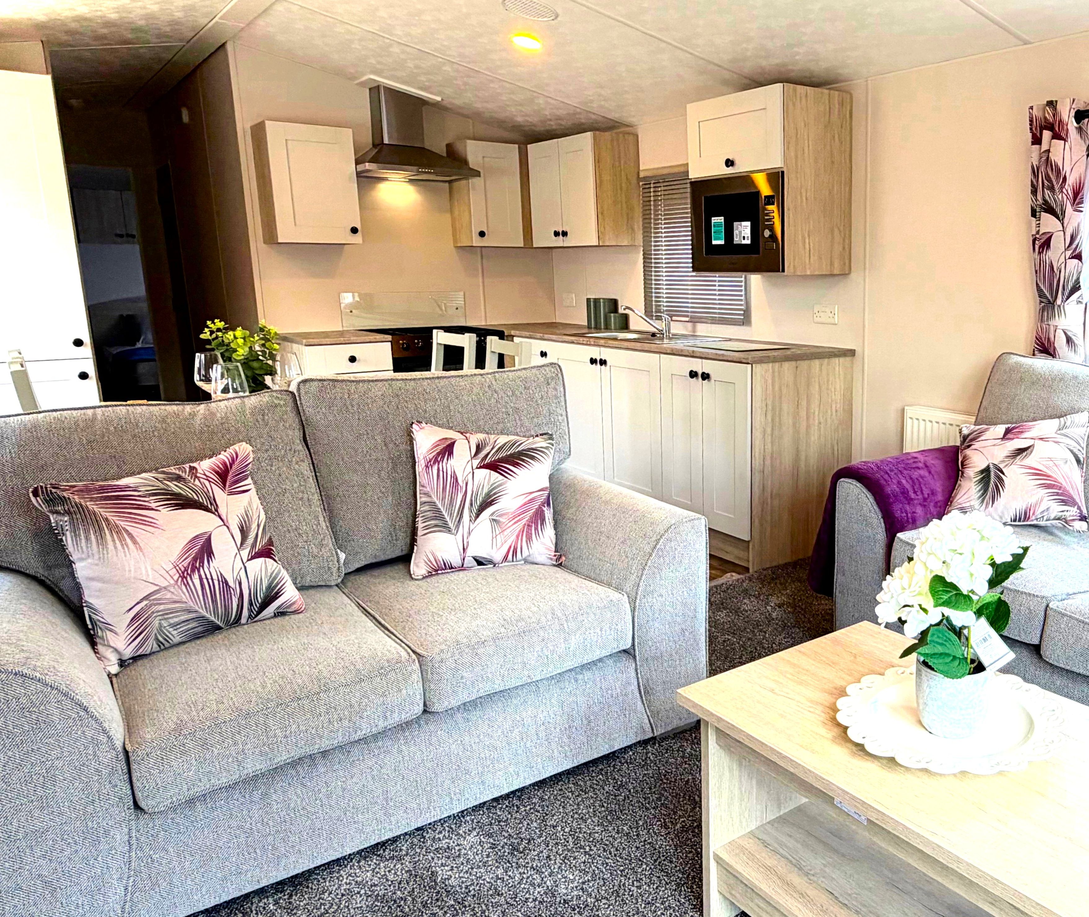 Modern open-plan lounge and kitchen with grey sofas, botanical cushions and light wood units