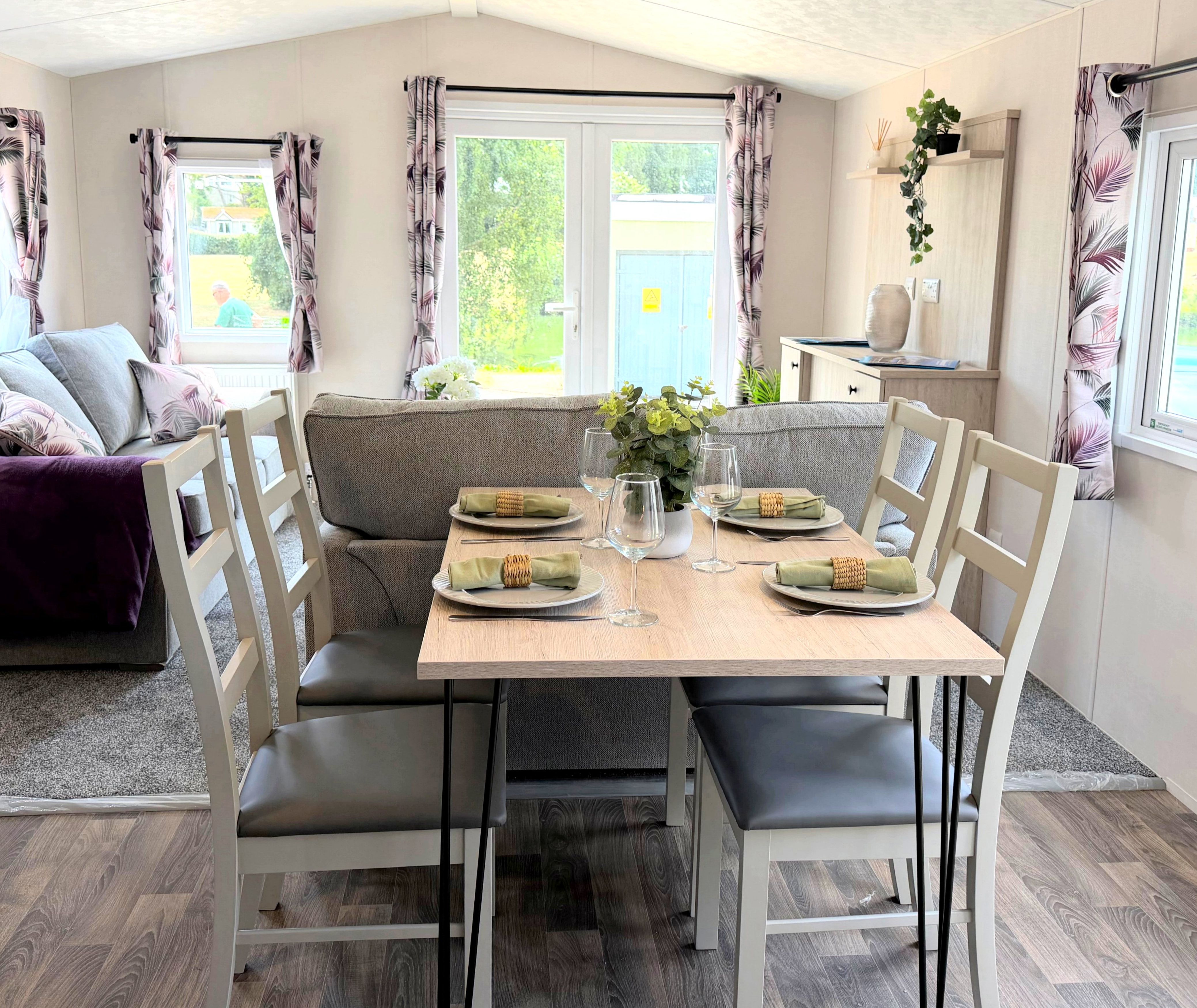 Dining table set for four in a bright open-plan living area with grey sofa and patterned curtains
