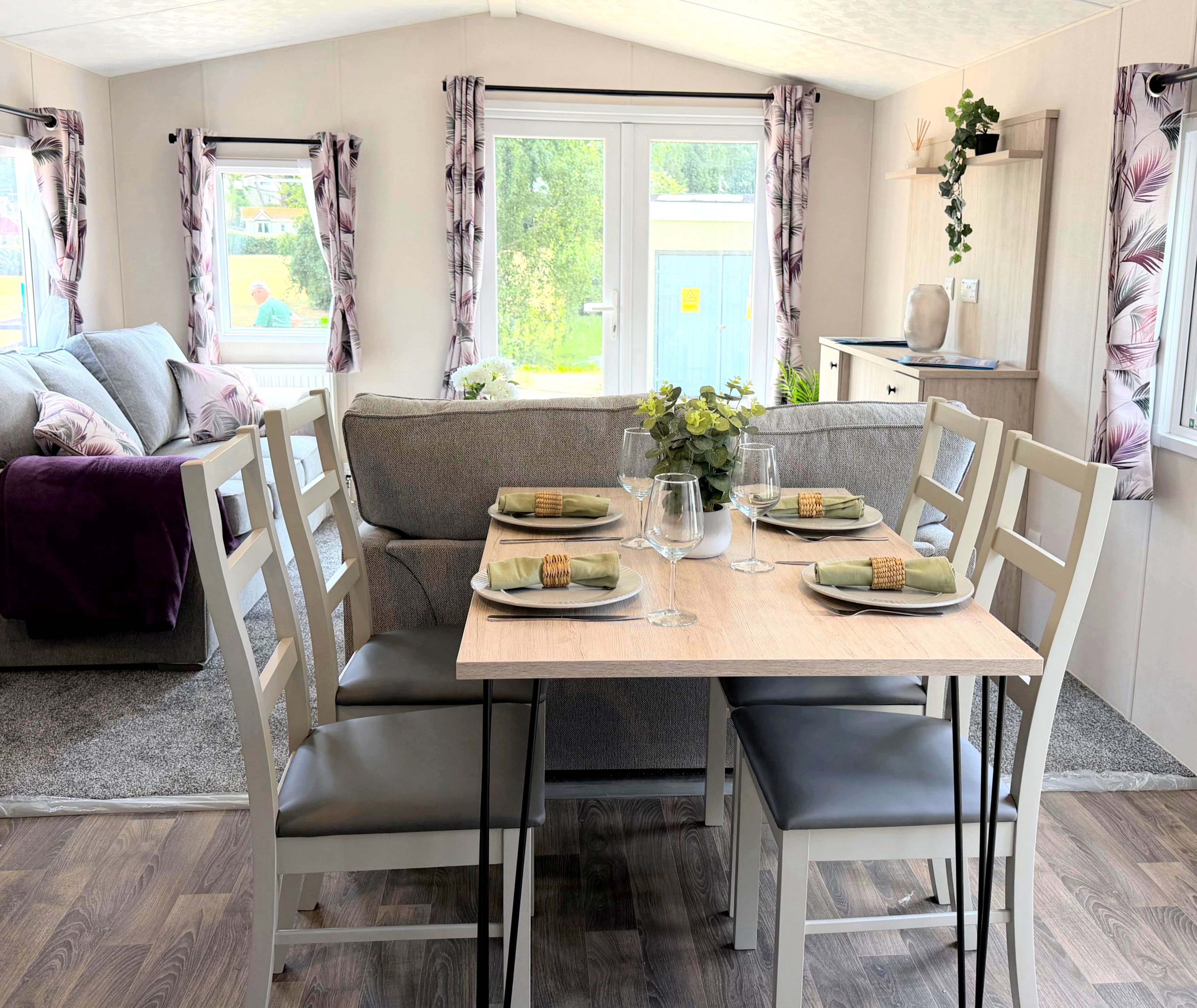 Dining table set for four in a bright open-plan living area with grey sofa and patterned curtains