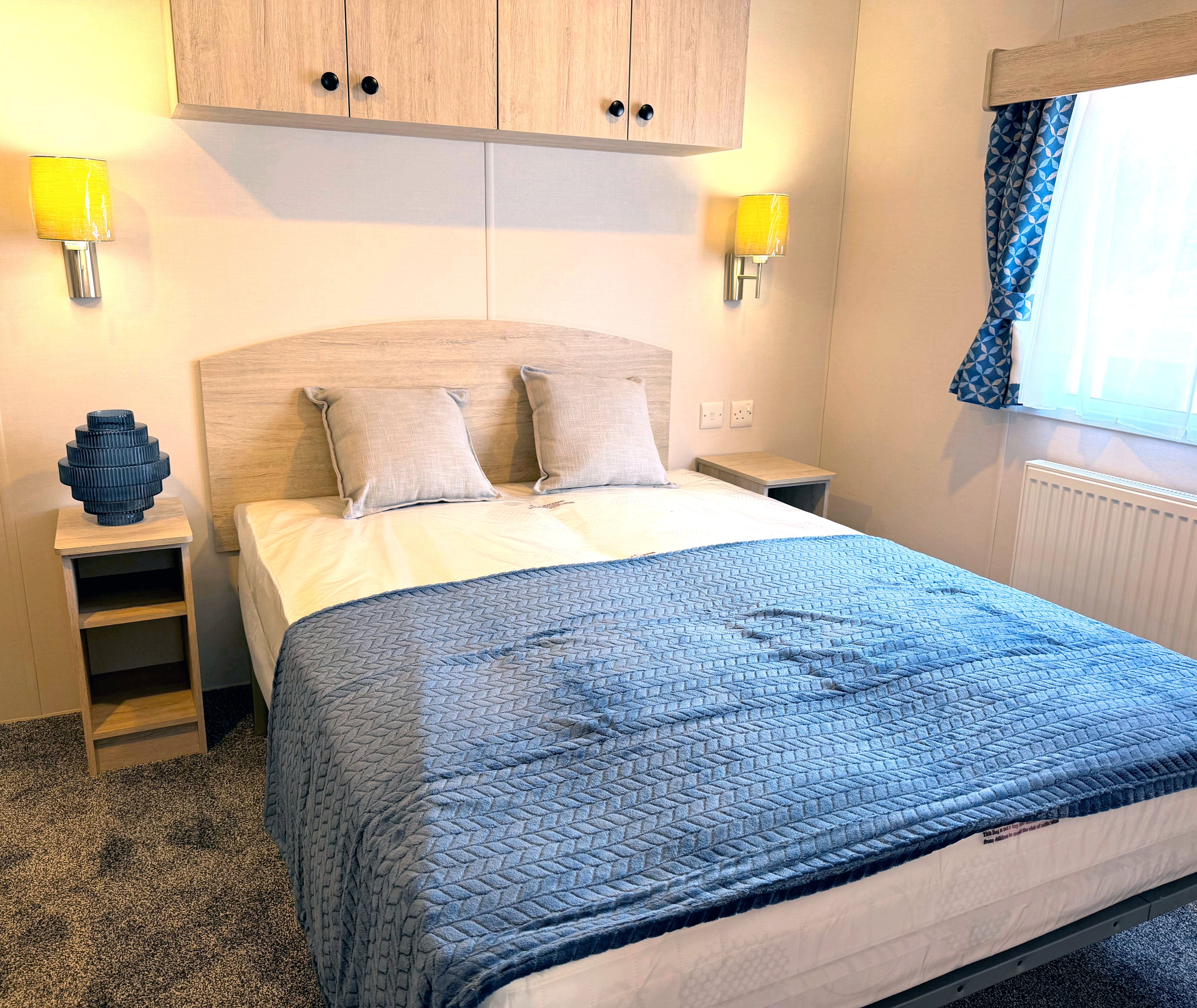 Master bedroom with double bed, blue quilted throw, wall lights and window with patterned curtains