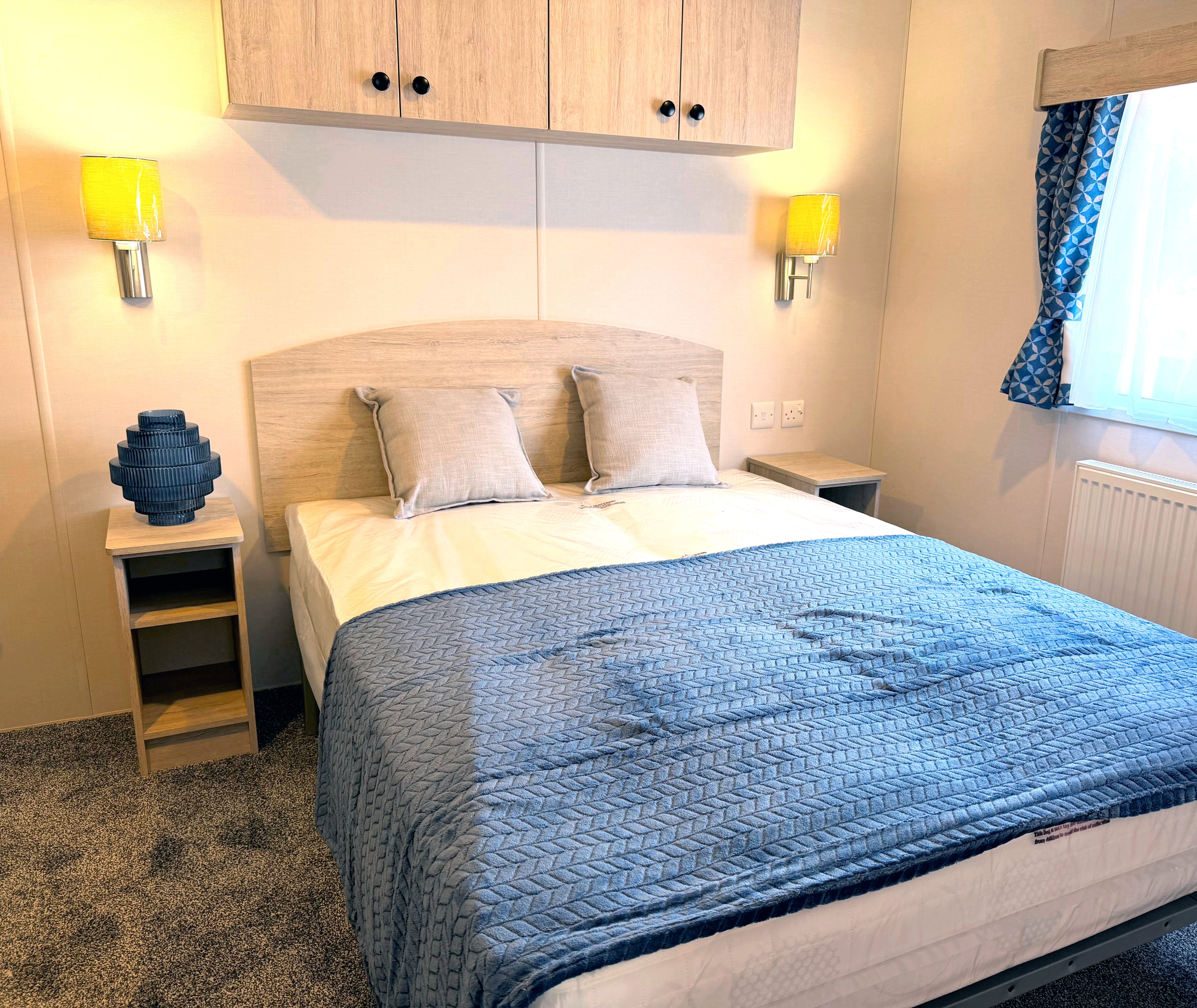 Master bedroom with double bed, blue quilted throw, wall lights and window with patterned curtains