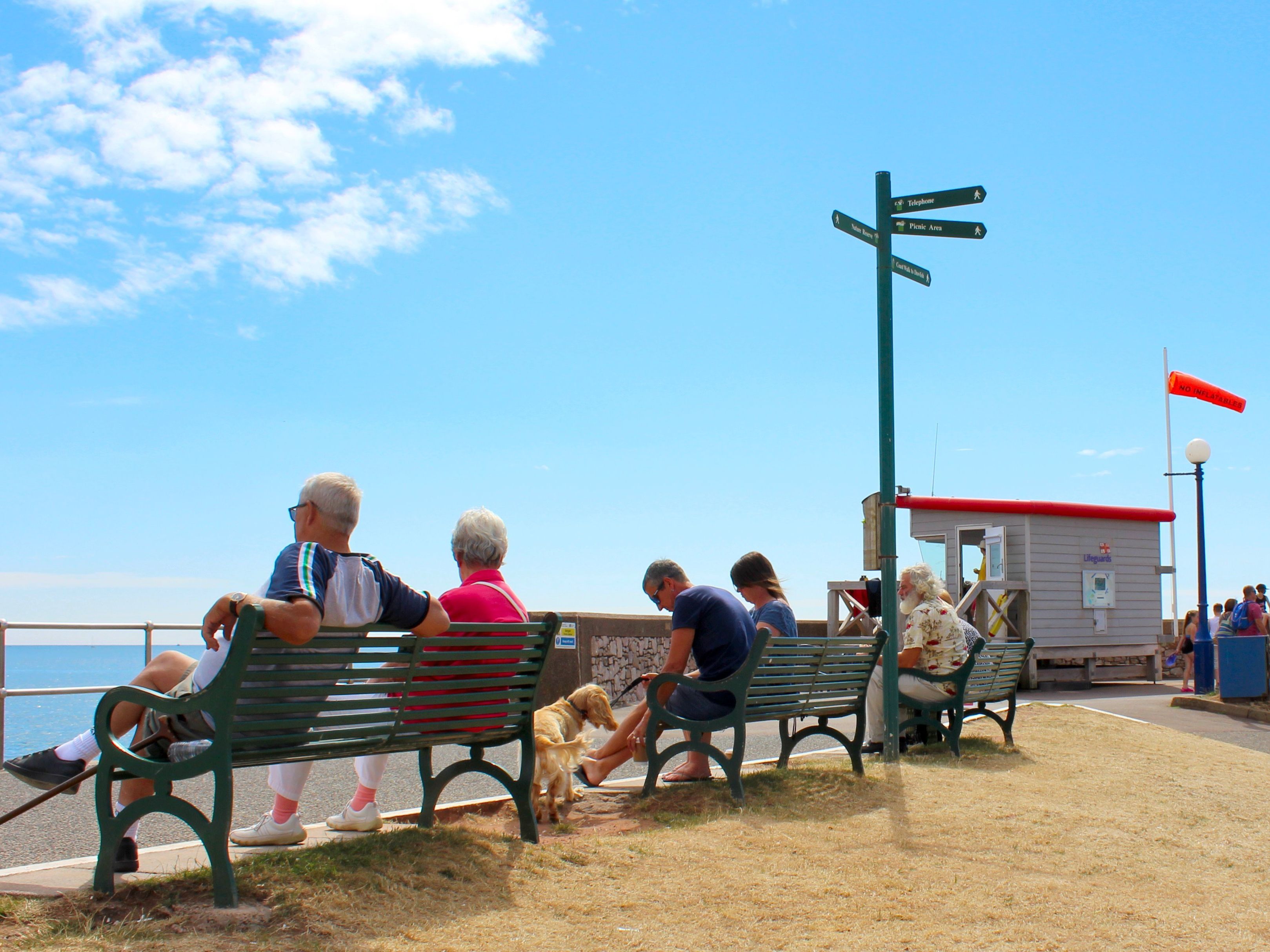 People sitting on benches along a seaside promenade near a lifeguard hut on a sunny day