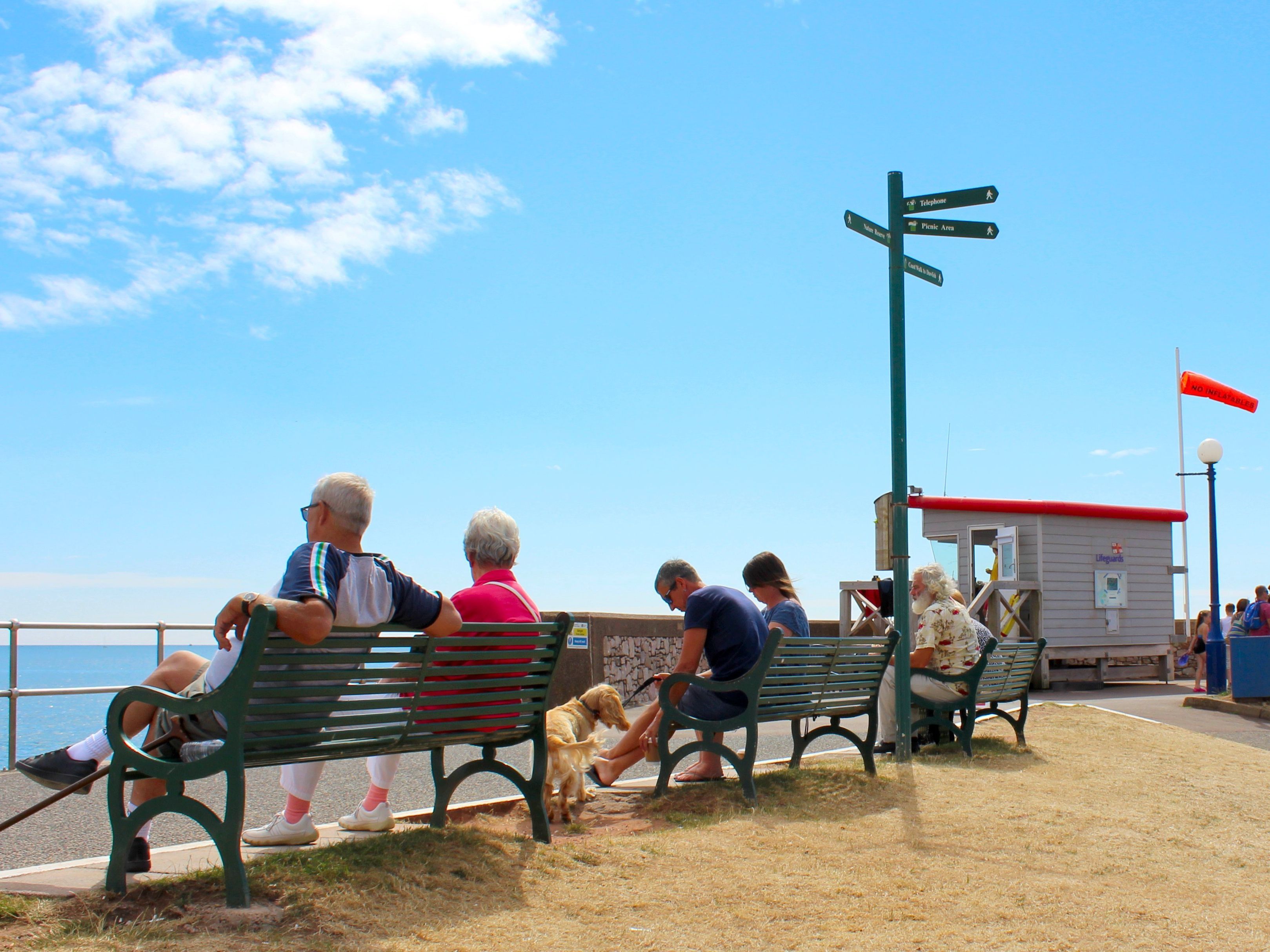 People sitting on benches along a seaside promenade near a lifeguard hut on a sunny day