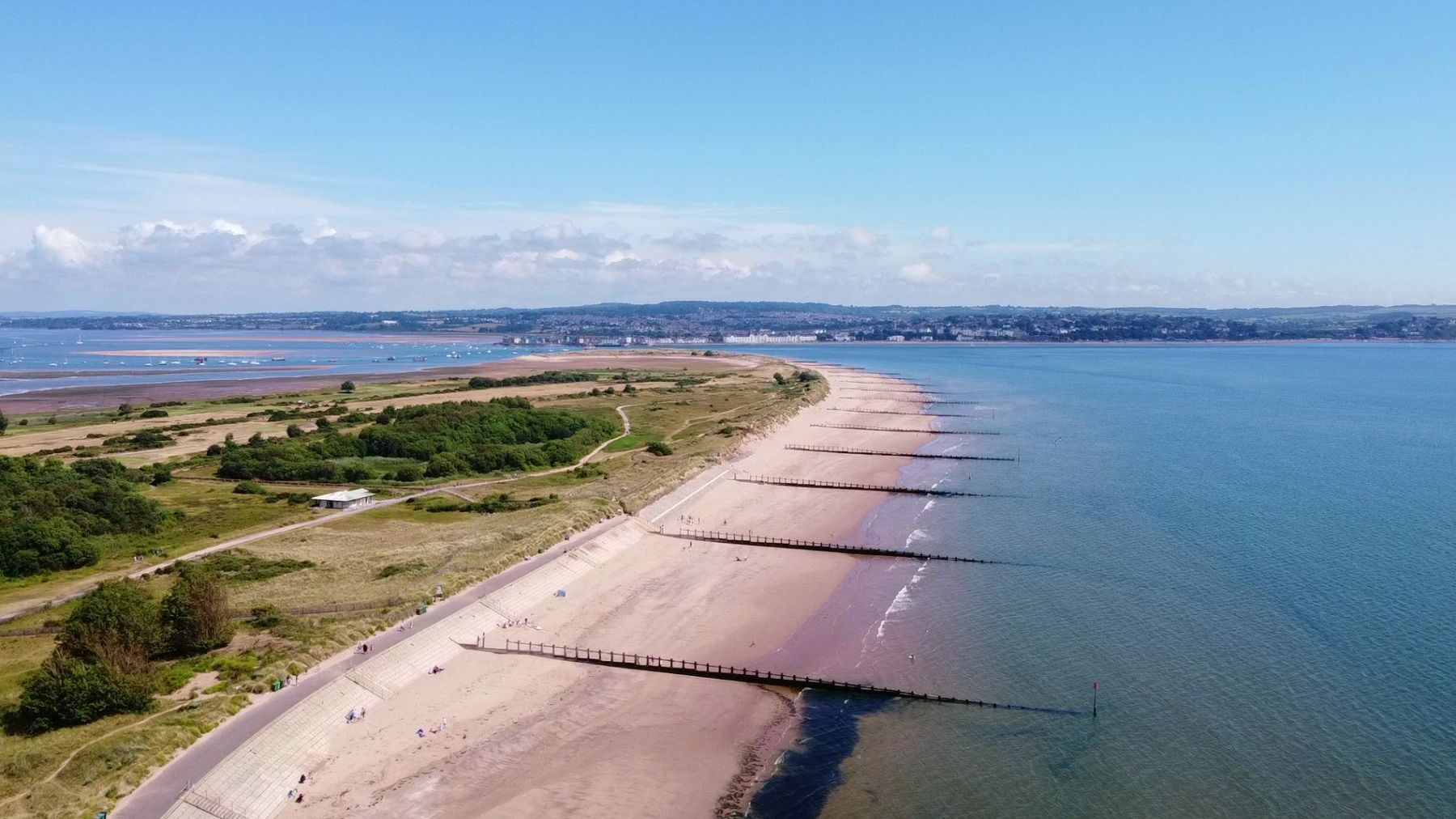 Aerial view of Dawlish Warren beach with sandy shoreline, sea groynes and dunes under a clear blue sky