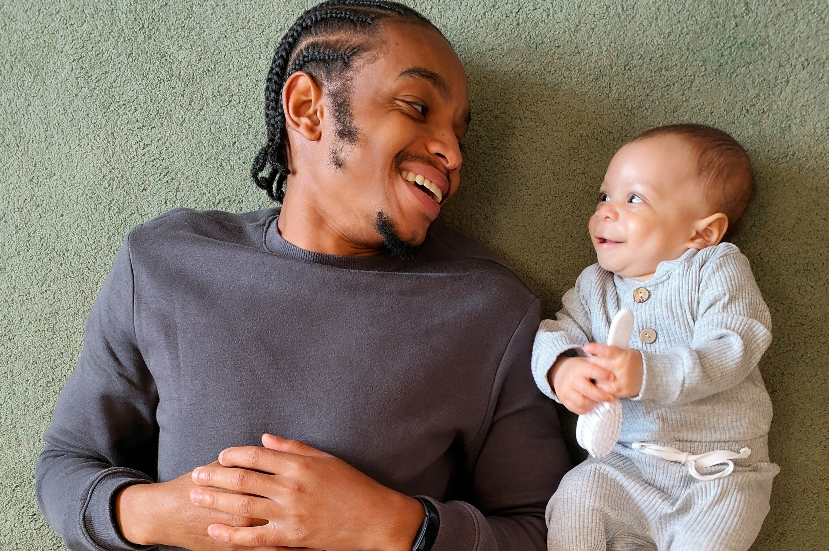 Father lying on a carpet smiling at his baby holding a spoon